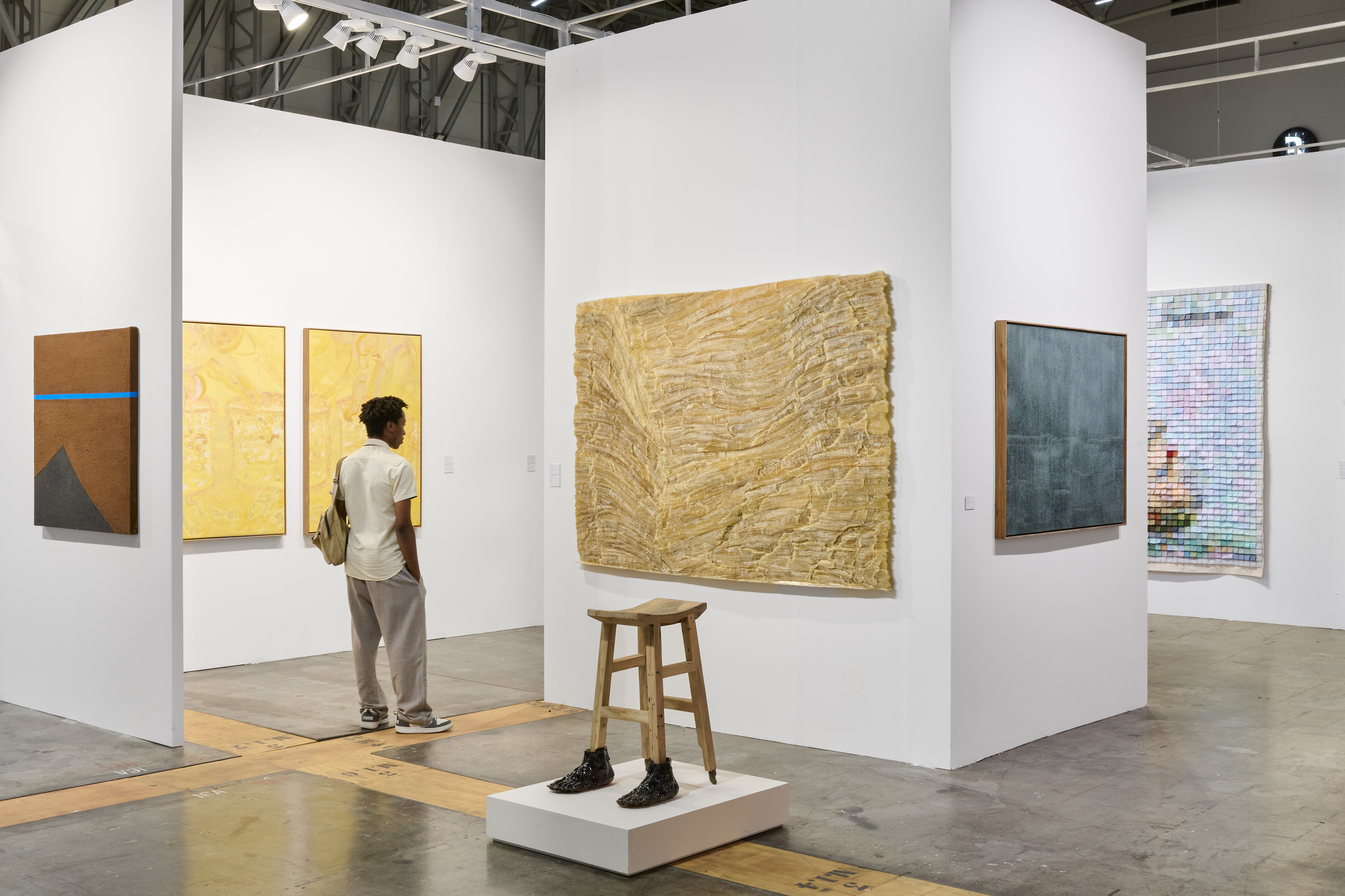 A person stands in a contemporary art gallery, observing diverse paintings and a wooden stool sculpture with shoes.