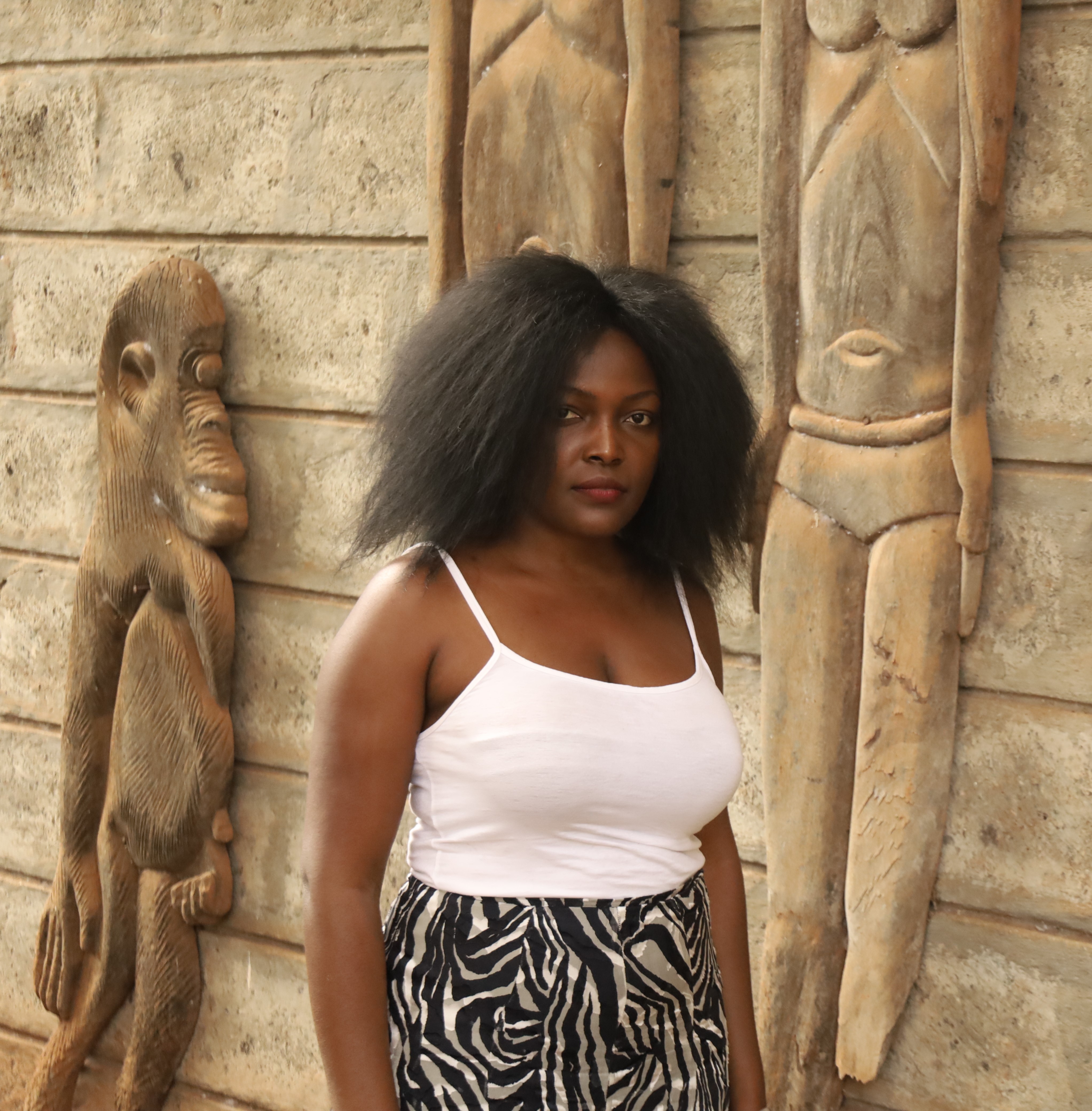 A woman with dark skin and voluminous black hair, wearing a white tank top and a black and white zebra print skirt, stands against a textured wall with wooden carvings.
