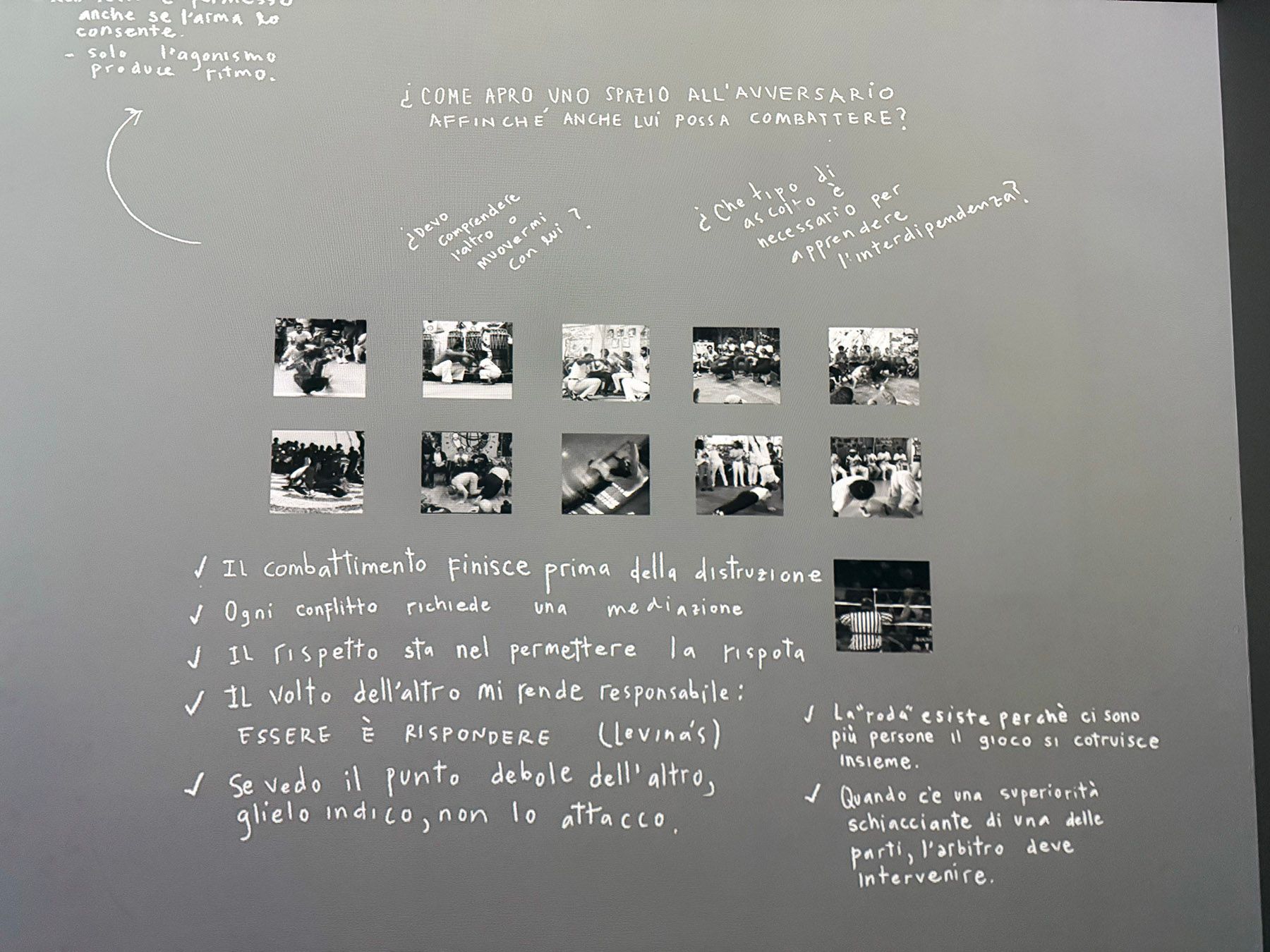 A gray exhibition wall with white Italian text about conflict, respect, and interdependence, alongside 13 black-and-white photos of martial arts, grappling, and a boxing ring.