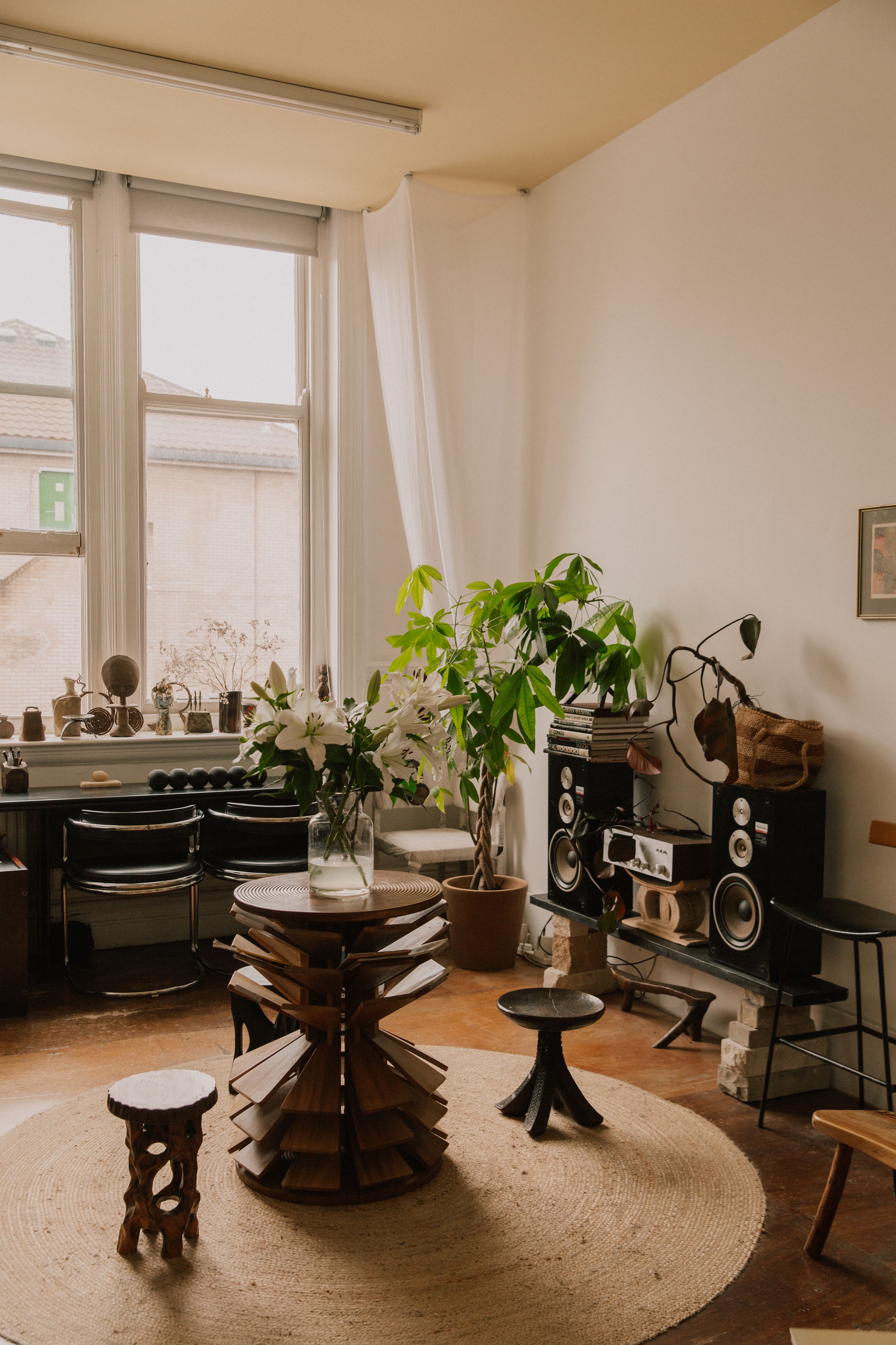 A bright, eclectic room with a large window, white lilies, a potted plant, unique wooden furniture, and vintage audio equipment on a round jute rug.