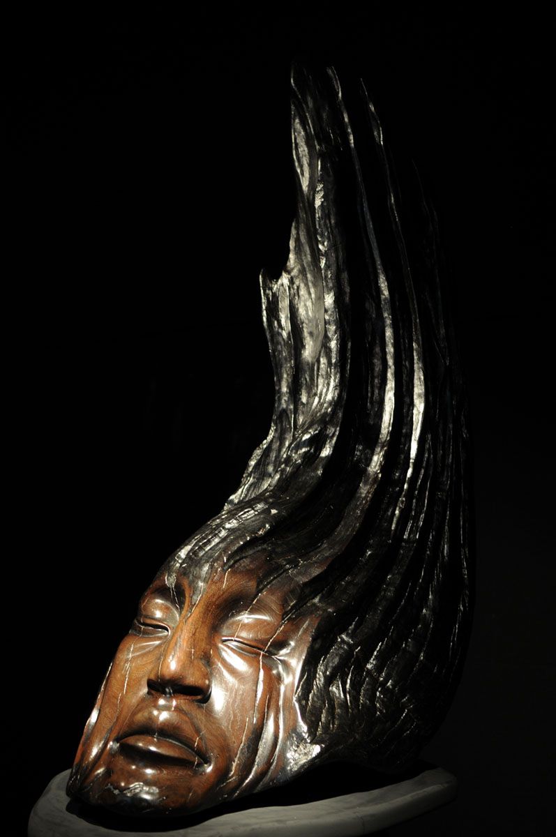 A dark wooden sculpture of a serene face with closed eyes, emerging from textured, upward-sweeping wood.