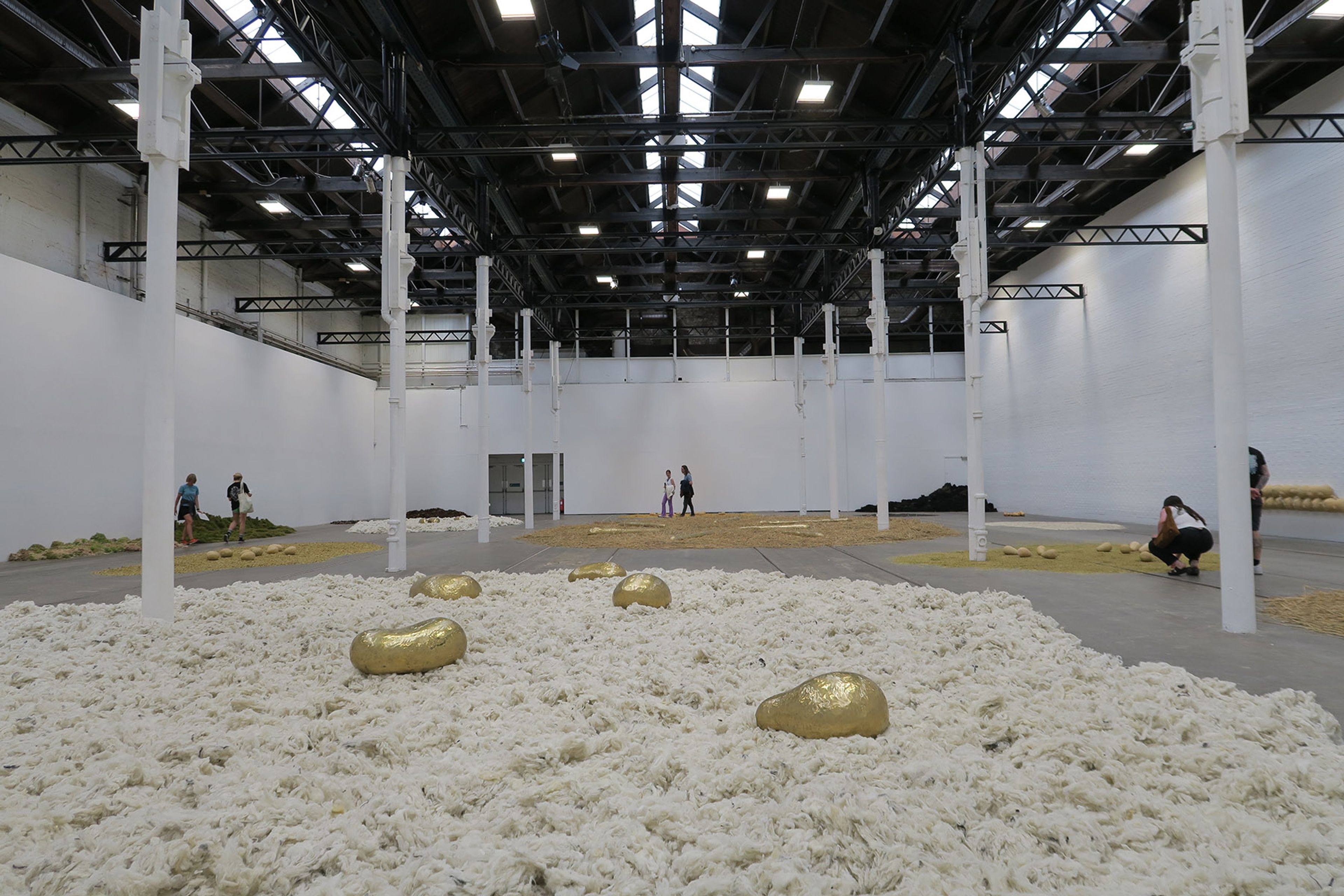 An art installation in a large gallery space, featuring a floor covered in white fibrous material, gold organic shapes, and other natural piles, with several visitors.
