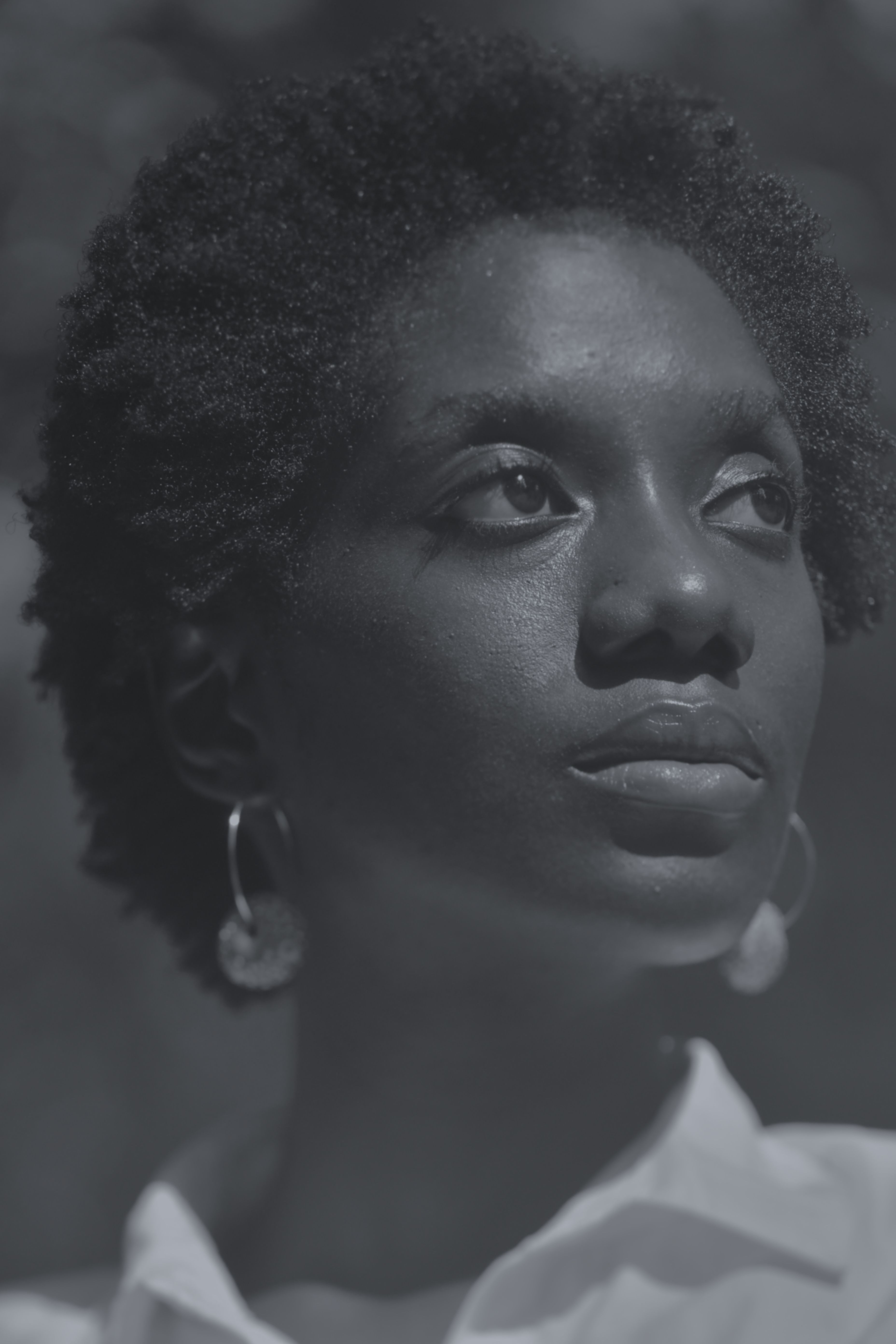 Close-up grayscale portrait of a woman with short curly hair, looking upwards with a thoughtful expression, wearing hoop earrings and a collared shirt.