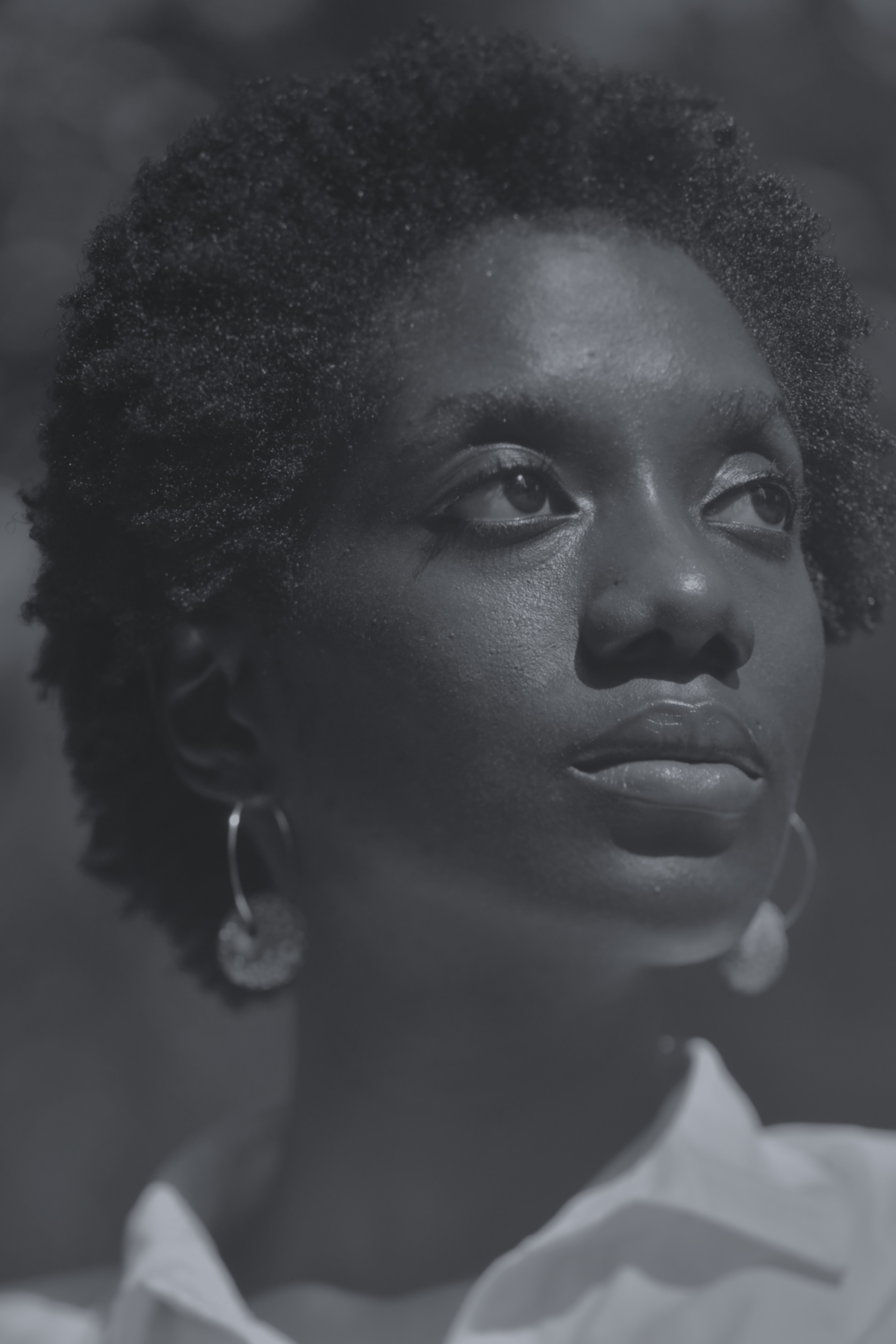 Close-up grayscale portrait of a woman with short curly hair, looking upwards with a thoughtful expression, wearing hoop earrings and a collared shirt.