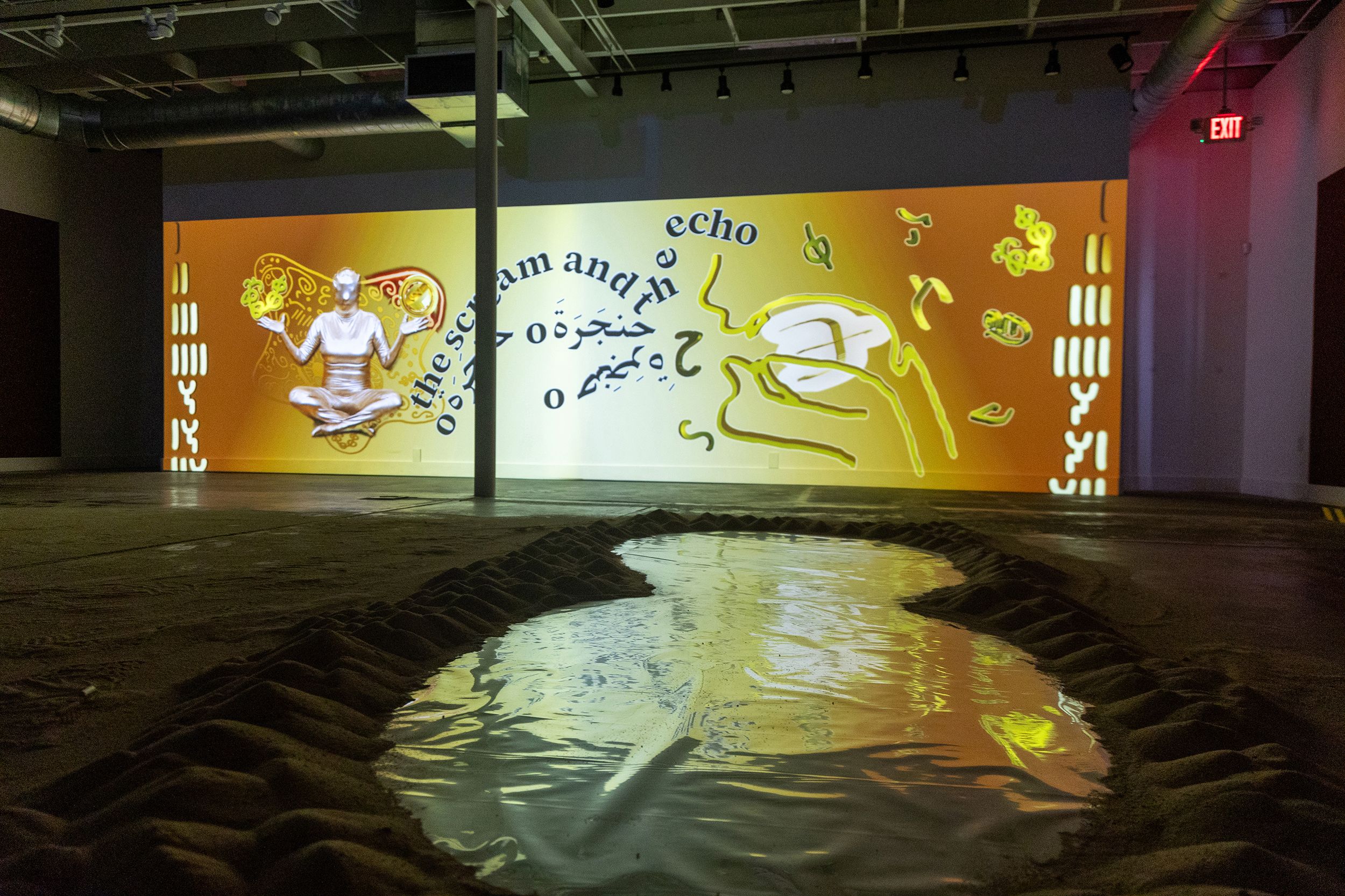 An art installation with a large orange wall projection showing a meditating figure, text, and abstract shapes, viewed over a winding pool of water surrounded by sand.