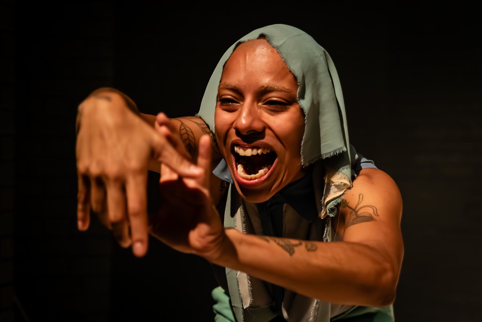 A person with dark skin and a light blue head covering screams with wide eyes and outstretched hands, illuminated against a dark background.