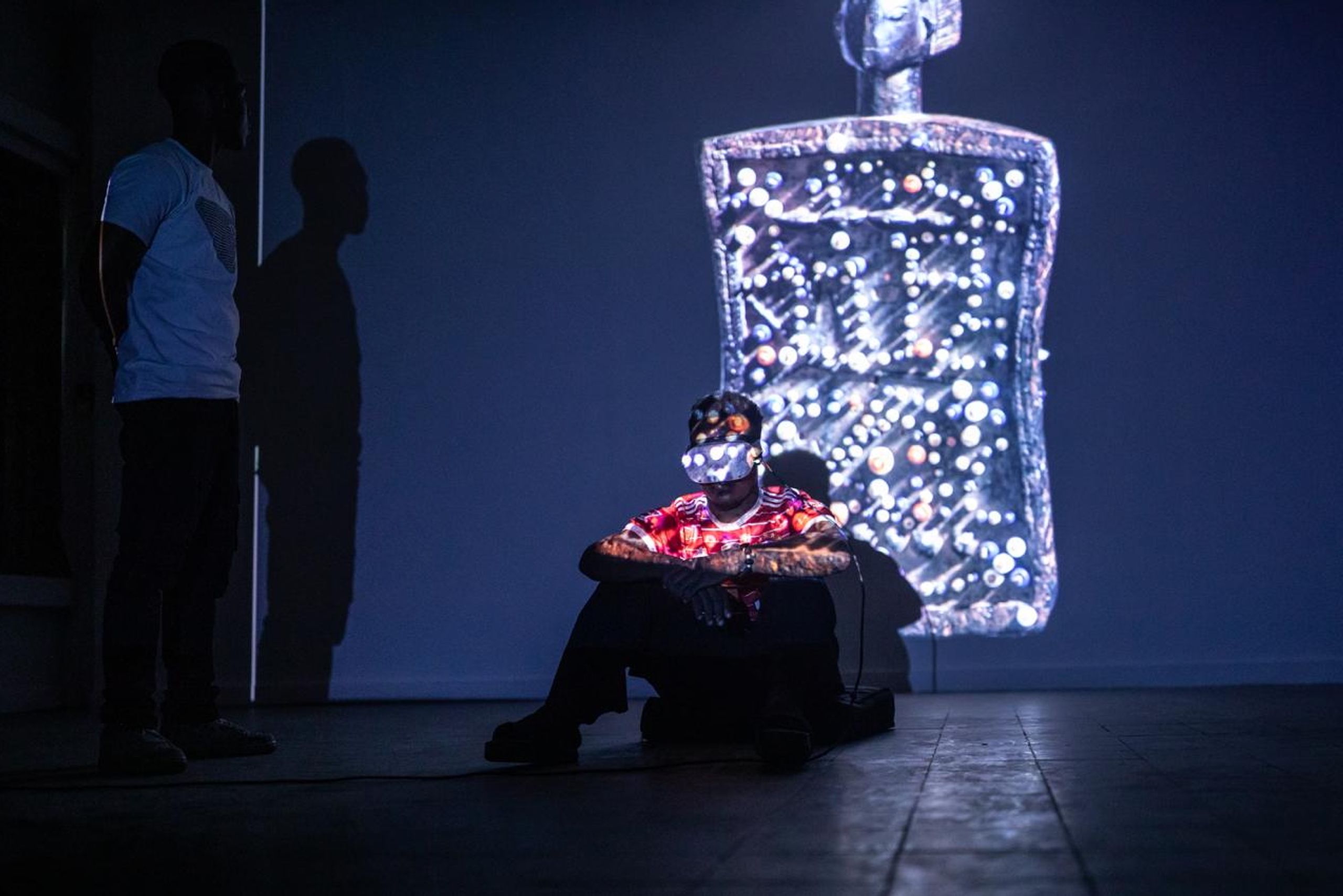 A person wearing a VR headset sits on the floor, illuminated by a large projection of a glowing, intricate digital figure on the wall, with another person standing in shadow.