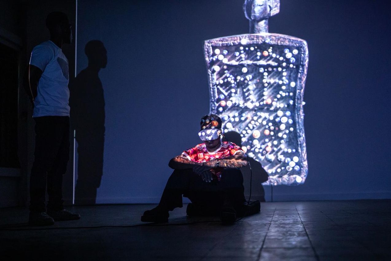 A person wearing a VR headset sits on the floor, illuminated by a large projection of a glowing, intricate digital figure on the wall, with another person standing in shadow.
