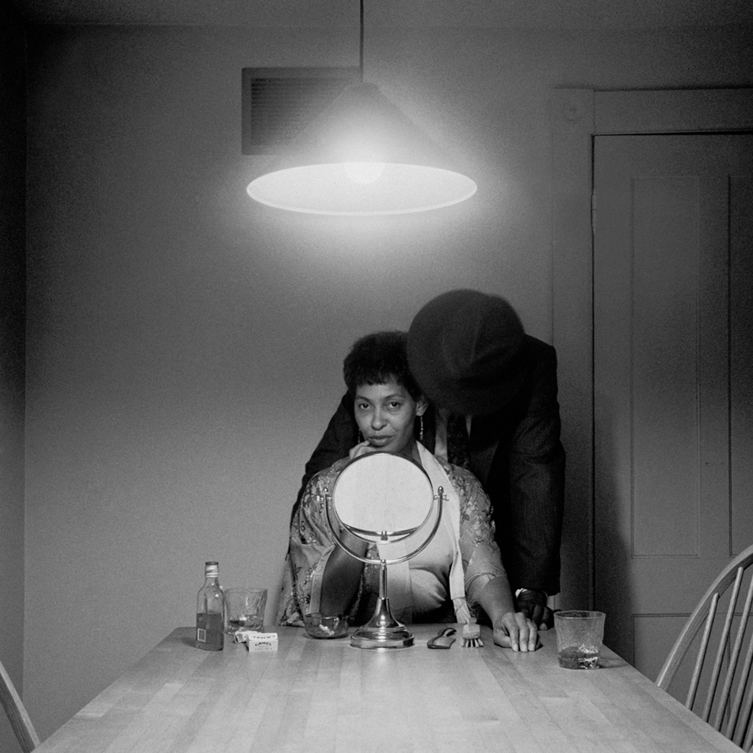 Black and white photograph of a woman looking into a table mirror, with a hat-clad man leaning over her shoulder, his face unseen. A bottle, glass, and cigarettes are on the wooden table under an overhead light.