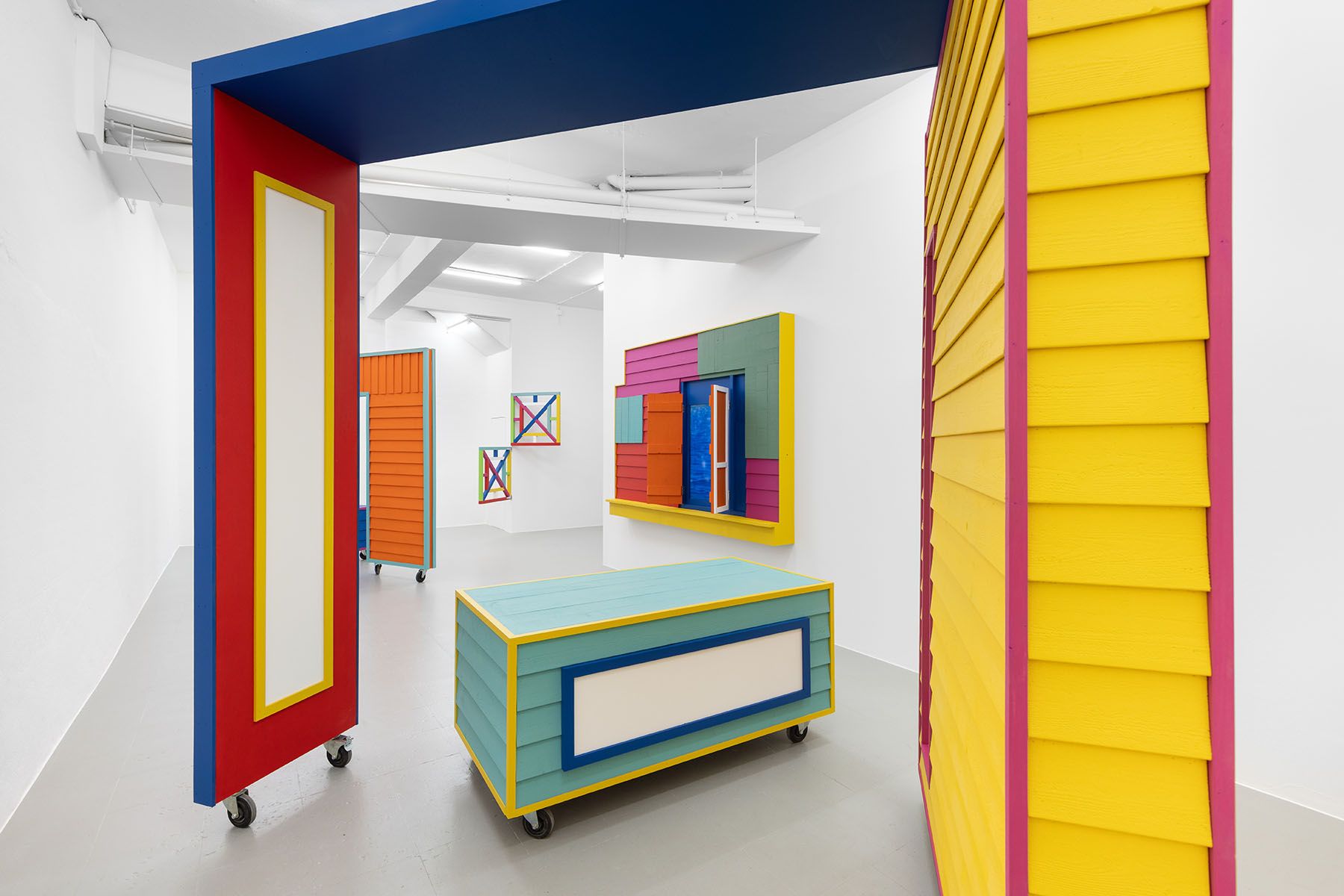 A vibrant art installation featuring modular, house-like structures with siding on wheels in a white gallery.