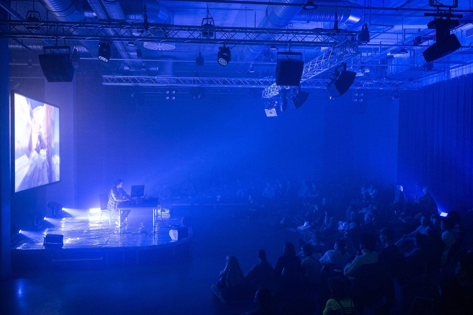 A DJ performs on a stage under blue spotlights, with a large screen, in front of a seated audience.
