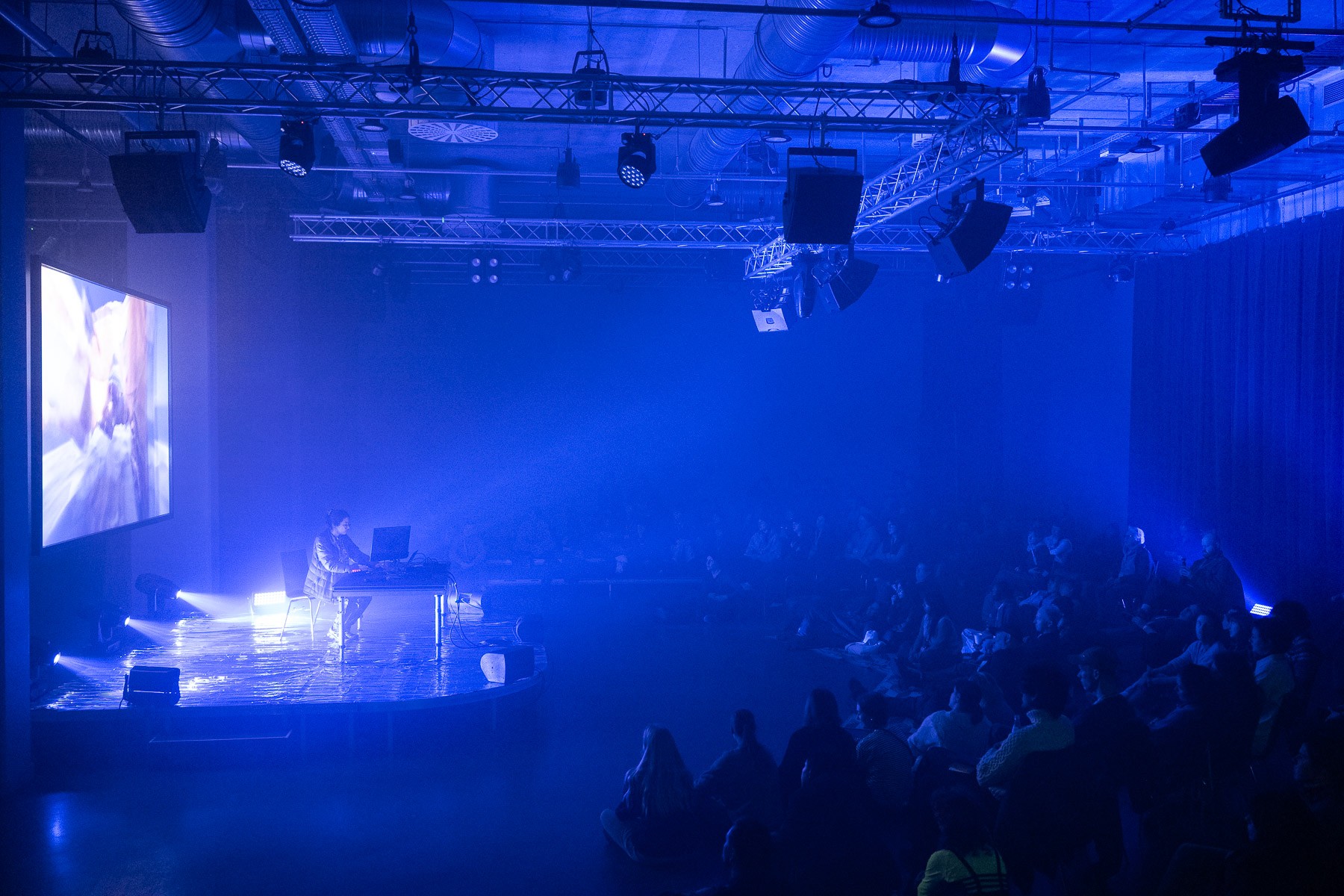 A DJ performs on a stage under blue spotlights, with a large screen, in front of a seated audience.