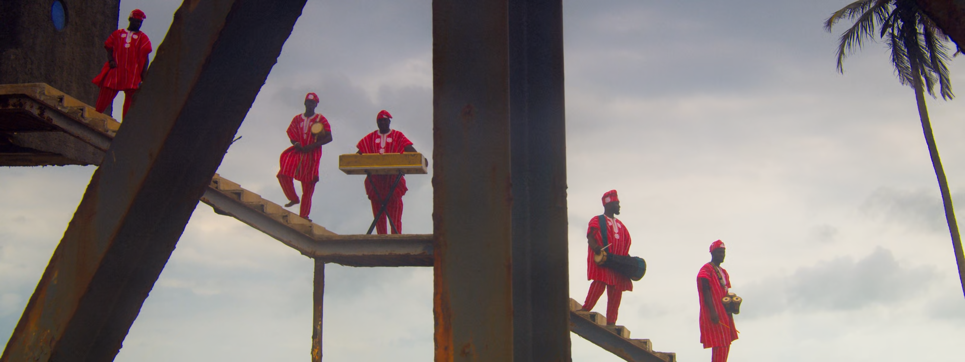 Six men in red traditional attire on a metal structure under a cloudy sky with a palm tree.