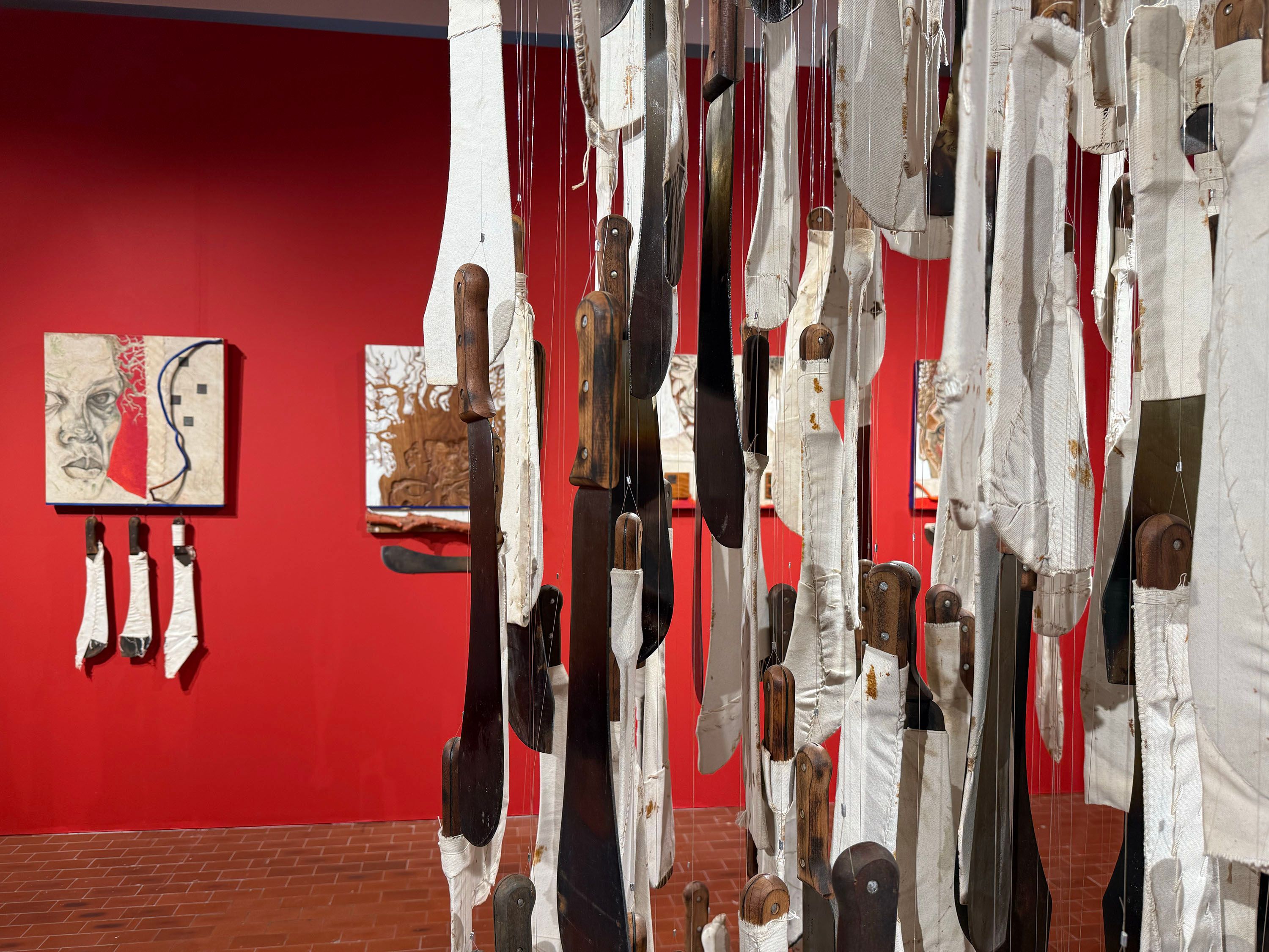An art gallery scene with a dense foreground installation of knives partially wrapped in white fabric, suspended before a bright red wall with other artworks, including a painting of a face.