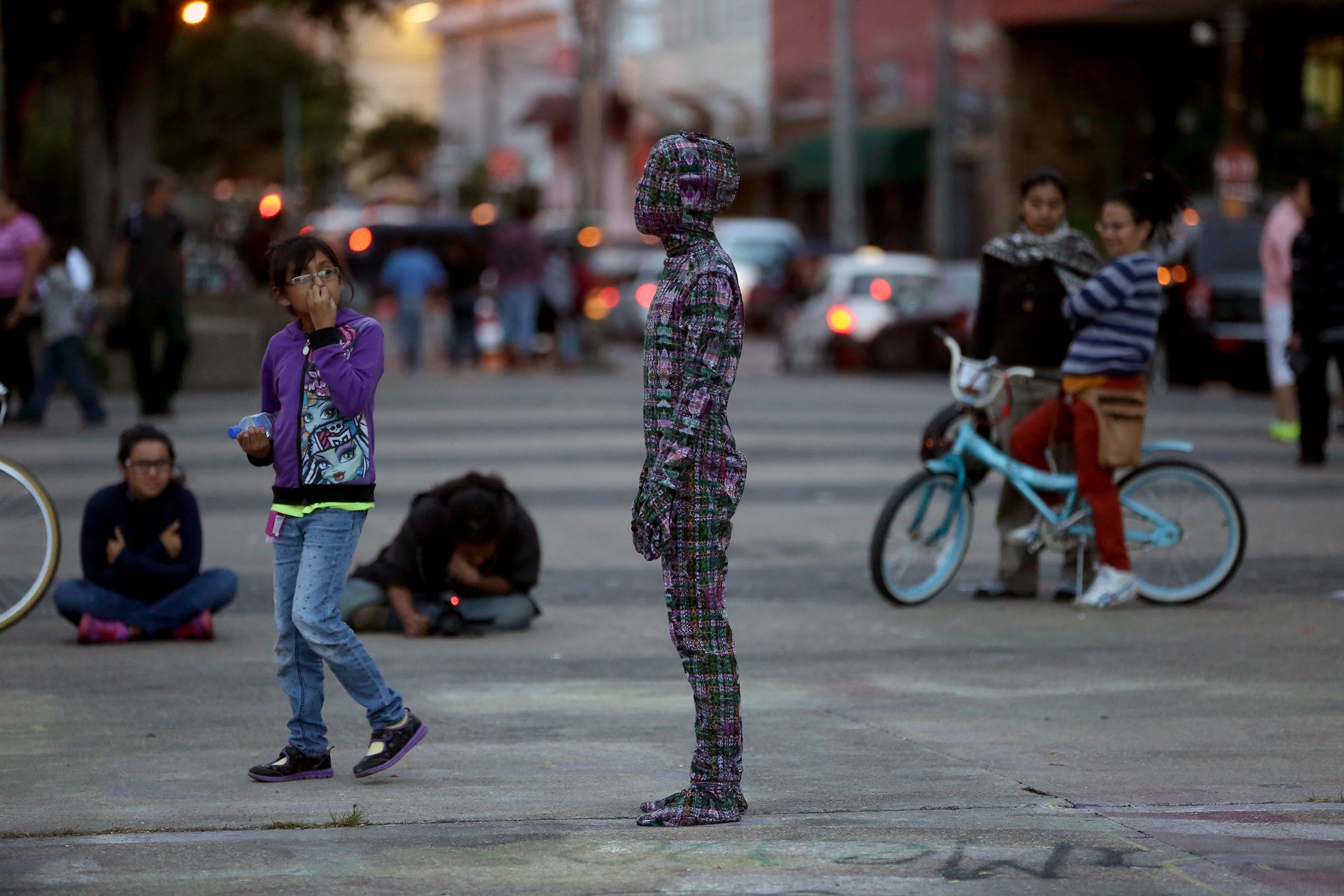 A street performer in a full plaid suit stands still in a public square, with onlookers and a blue bicycle nearby.