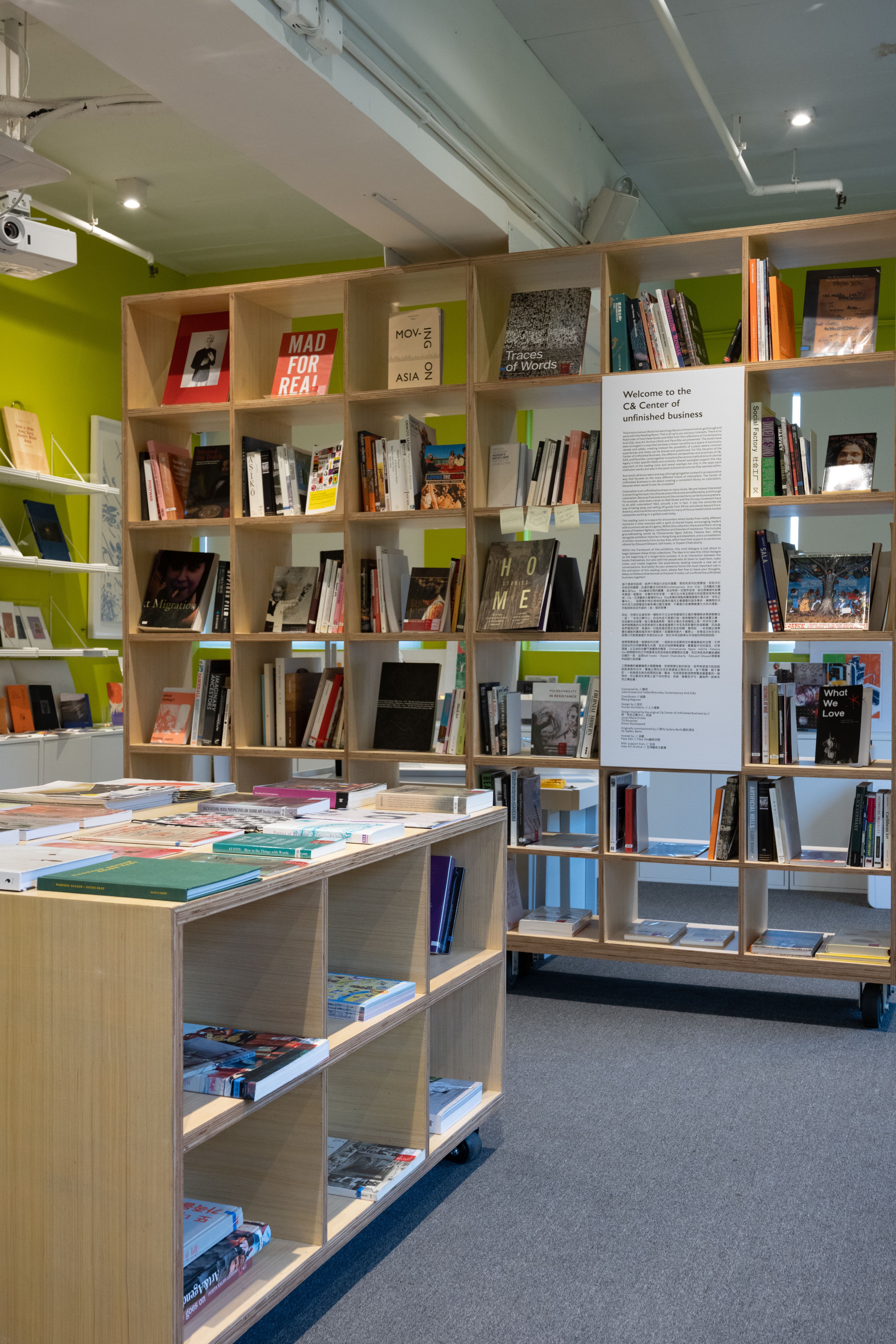A bright room with lime green walls, featuring light wood bookshelves and display tables filled with books, with a sign reading "Welcome to the C&R Center of unfinished business."