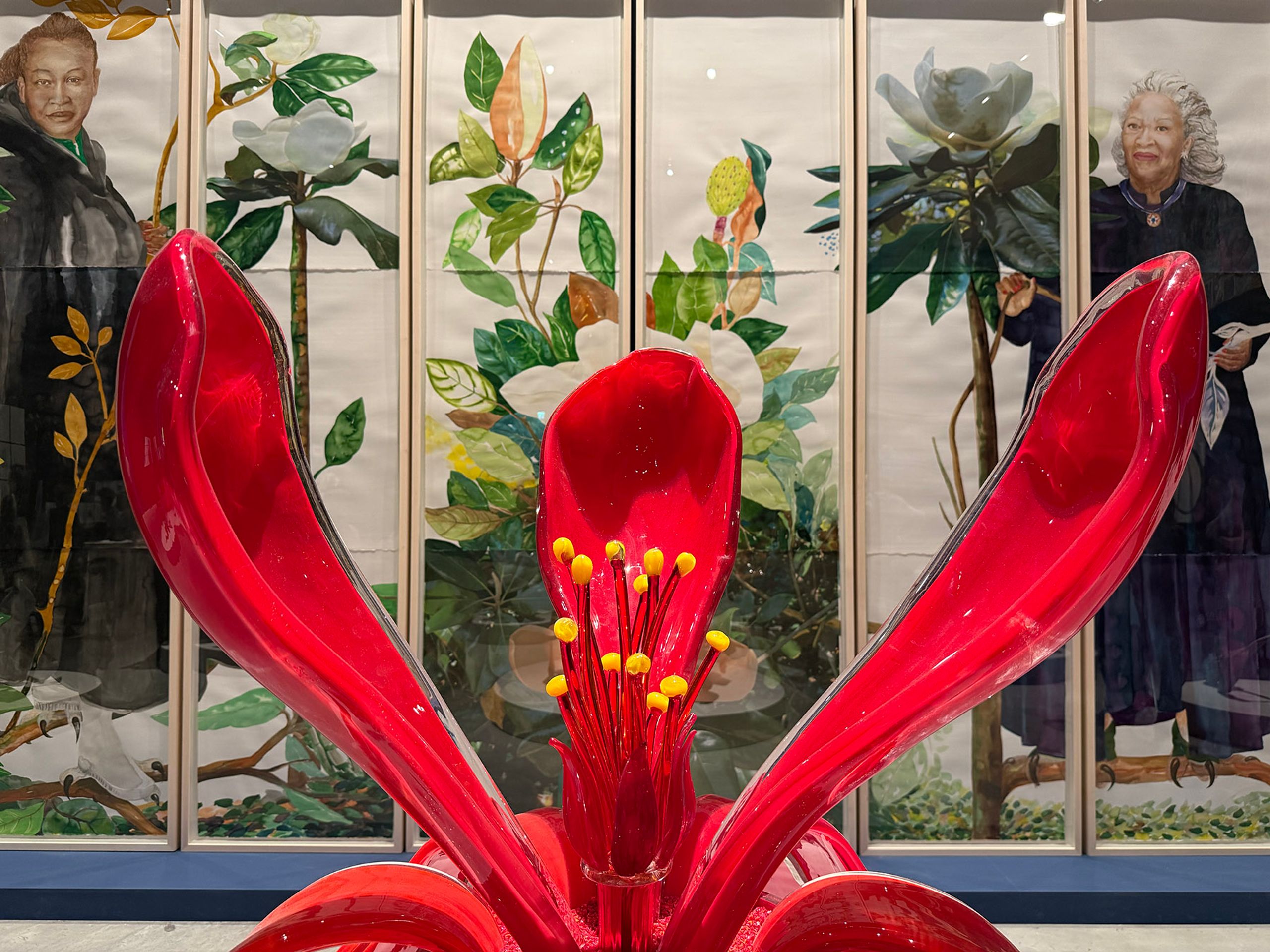 A vibrant red flower sculpture with prominent yellow stamens in the foreground. Behind it, multi-panel artwork depicts portraits of a woman with dark hair and a woman with white hair, interspersed with botanical illustrations of magnolias.