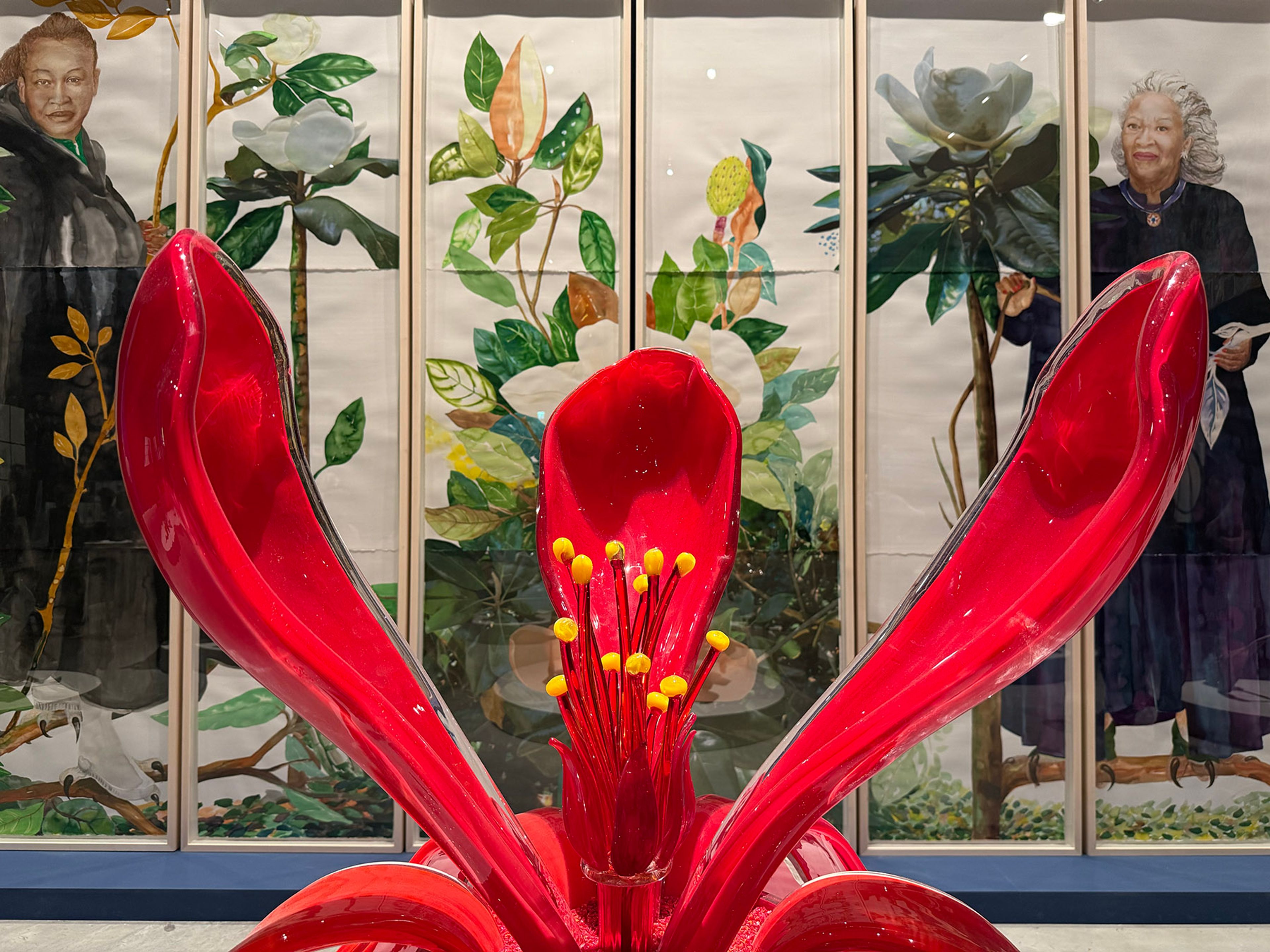 A vibrant red flower sculpture with prominent yellow stamens in the foreground. Behind it, multi-panel artwork depicts portraits of a woman with dark hair and a woman with white hair, interspersed with botanical illustrations of magnolias.