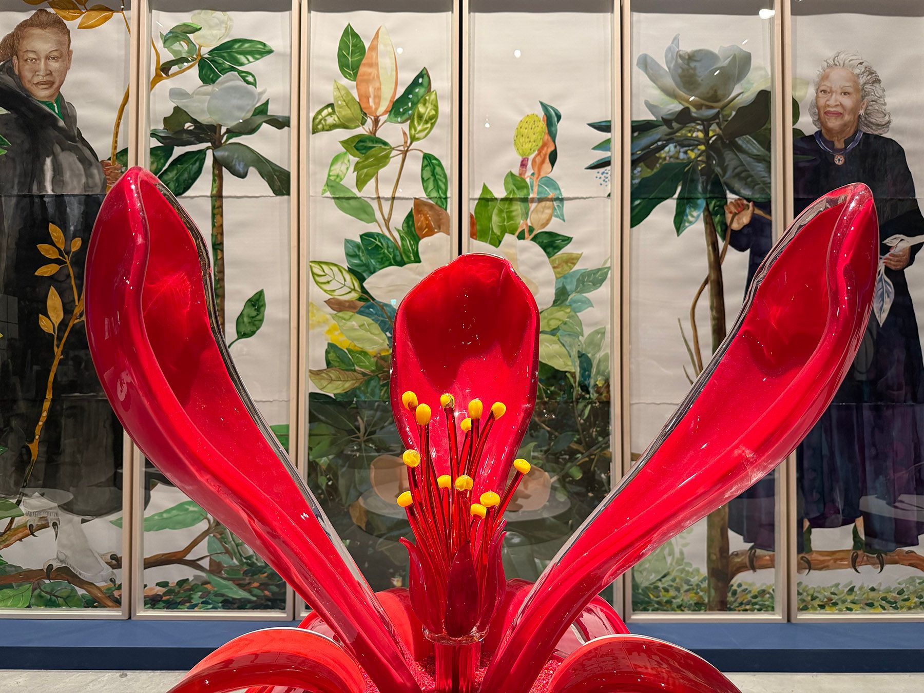 A vibrant red flower sculpture with prominent yellow stamens in the foreground. Behind it, multi-panel artwork depicts portraits of a woman with dark hair and a woman with white hair, interspersed with botanical illustrations of magnolias.