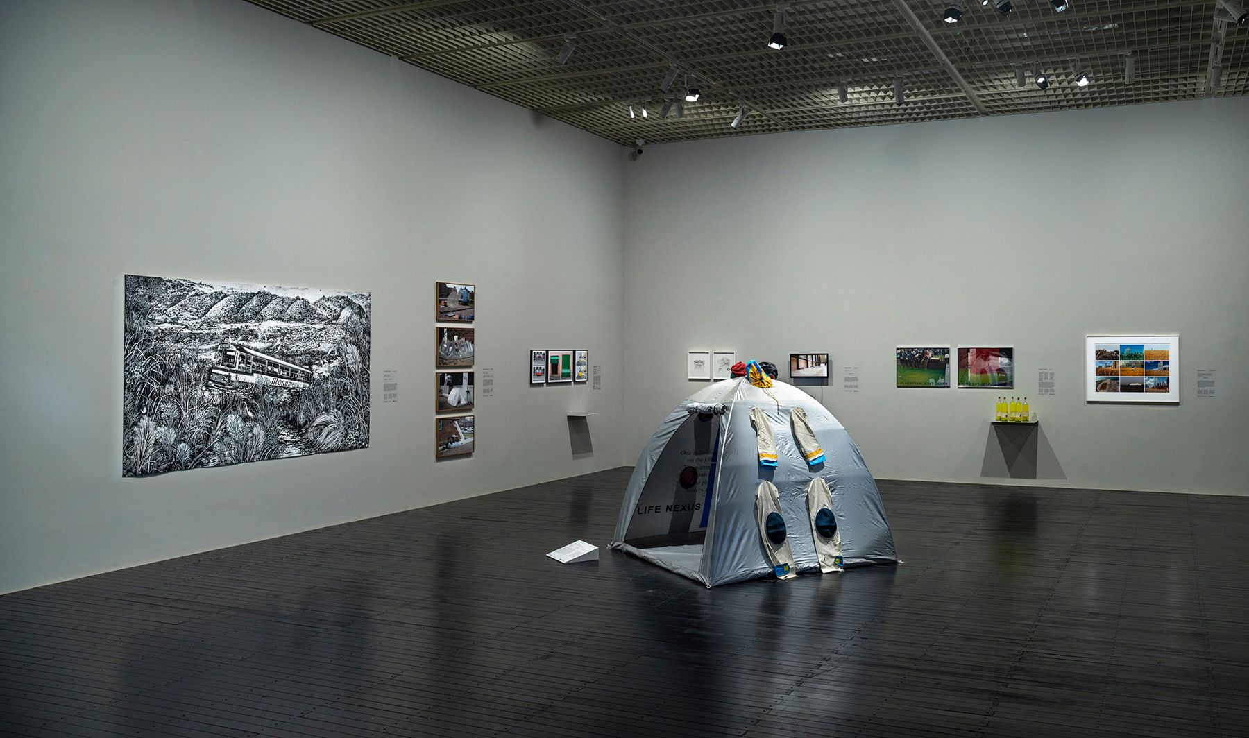 An art gallery installation featuring a large black and white landscape drawing, multiple framed prints, and a gray tent with two suits hanging from it.