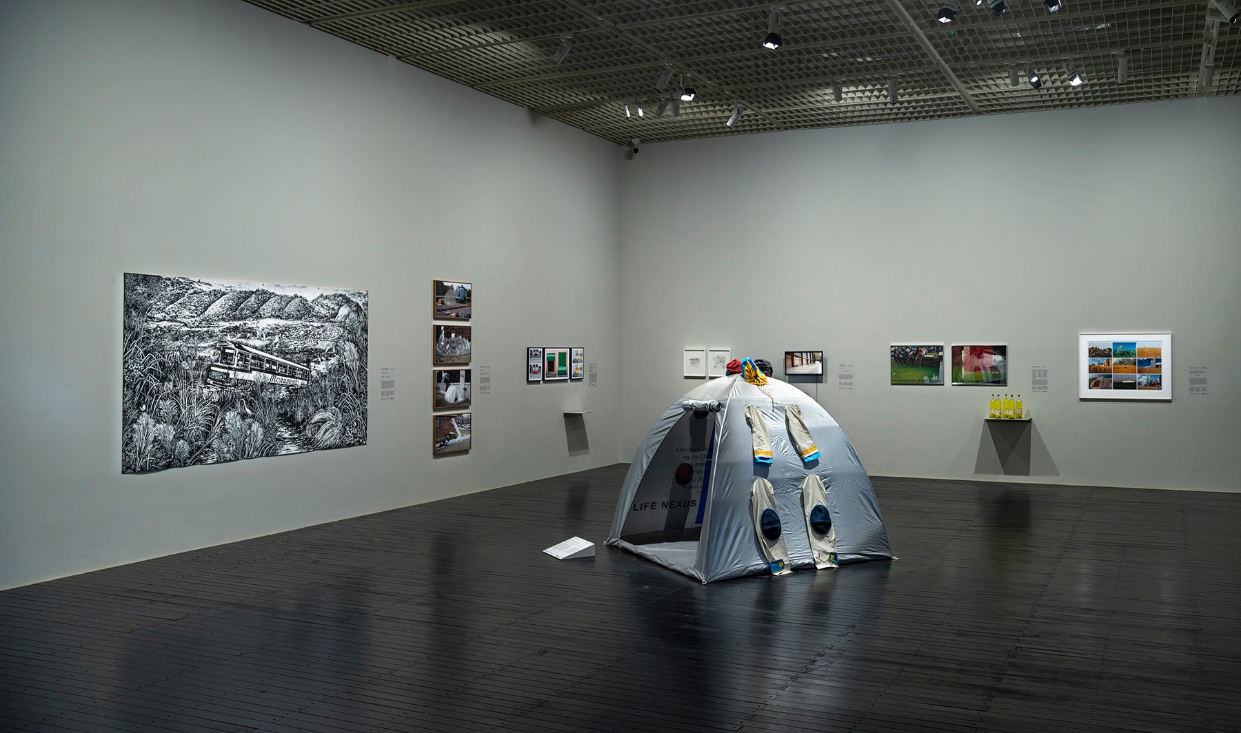 An art gallery installation featuring a large black and white landscape drawing, multiple framed prints, and a gray tent with two suits hanging from it.