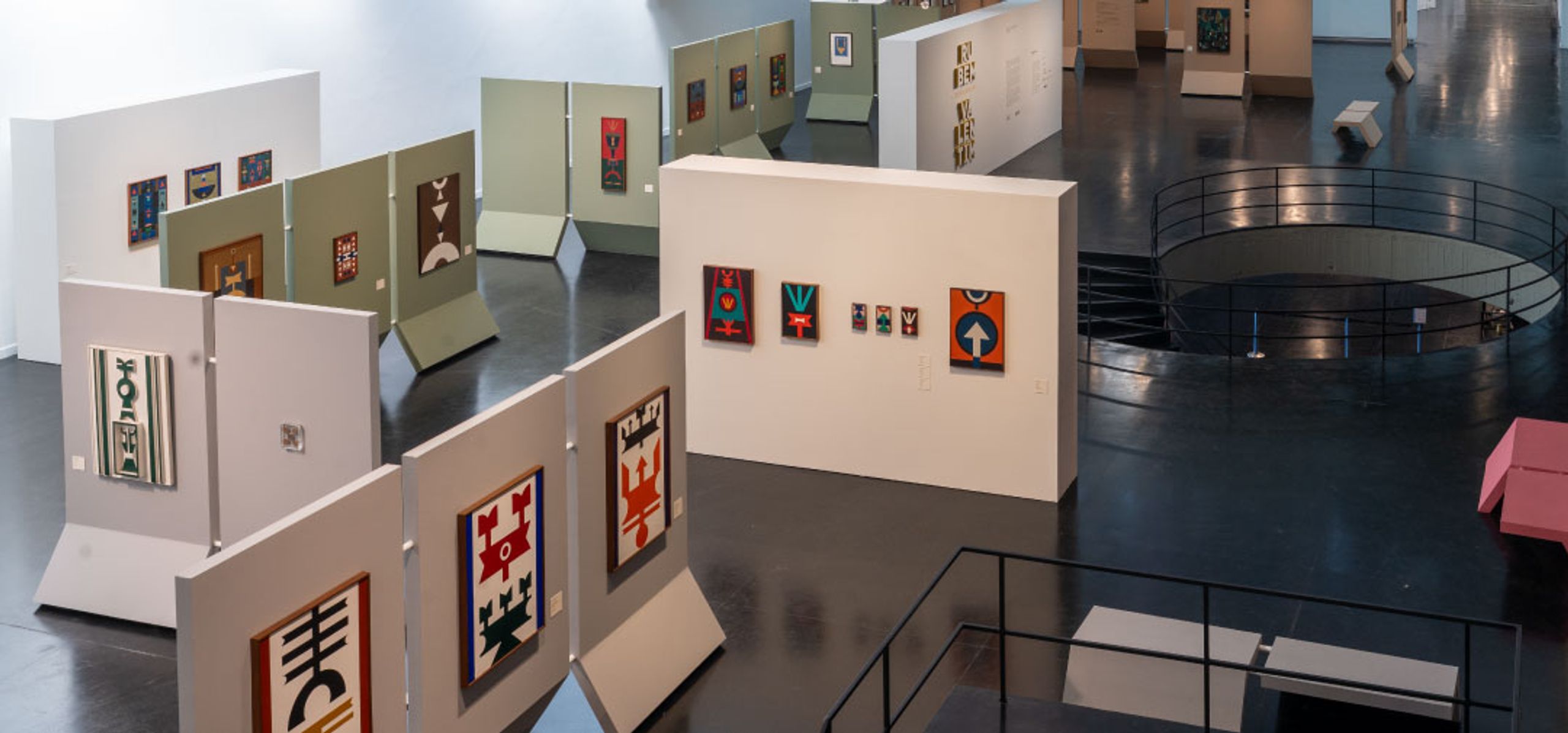 Art gallery displaying colorful geometric textile artworks on various white and light green panels, with a spiral staircase.