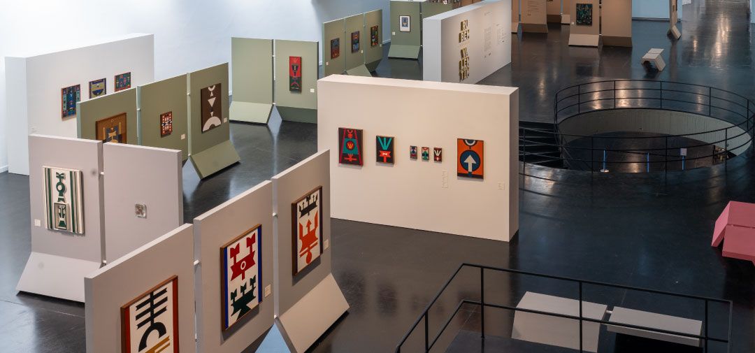 Art gallery displaying colorful geometric textile artworks on various white and light green panels, with a spiral staircase.