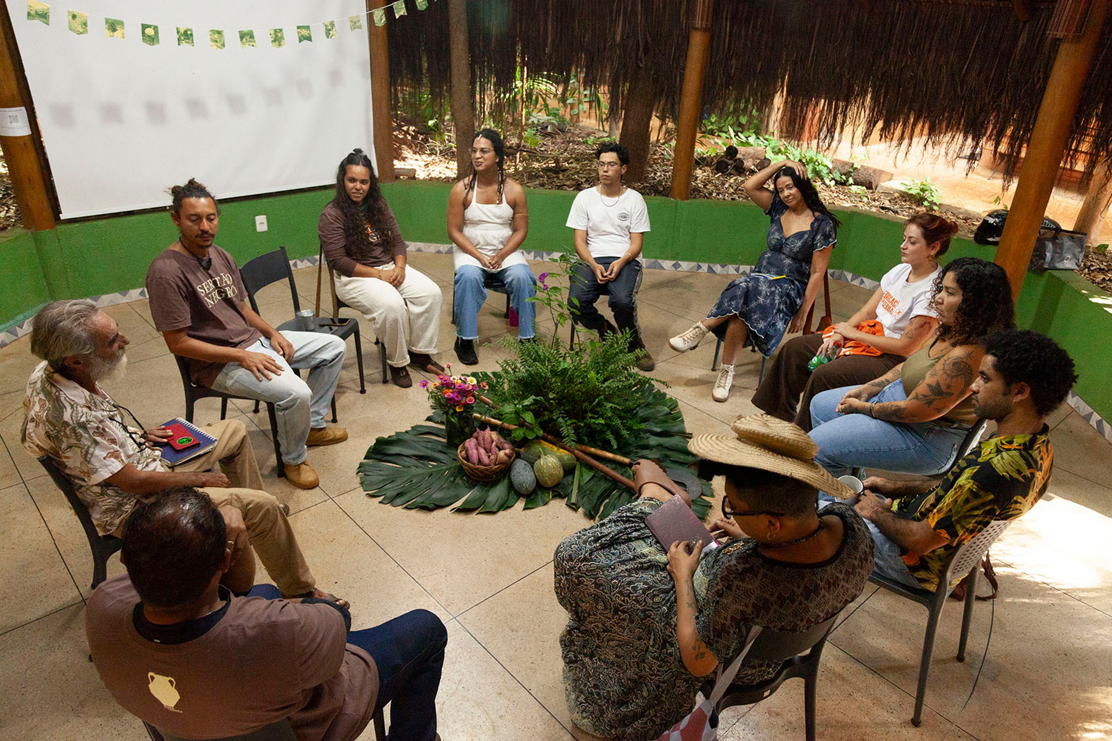 A diverse group of people sits in a circle around an arrangement of leaves, fruits, and roots.