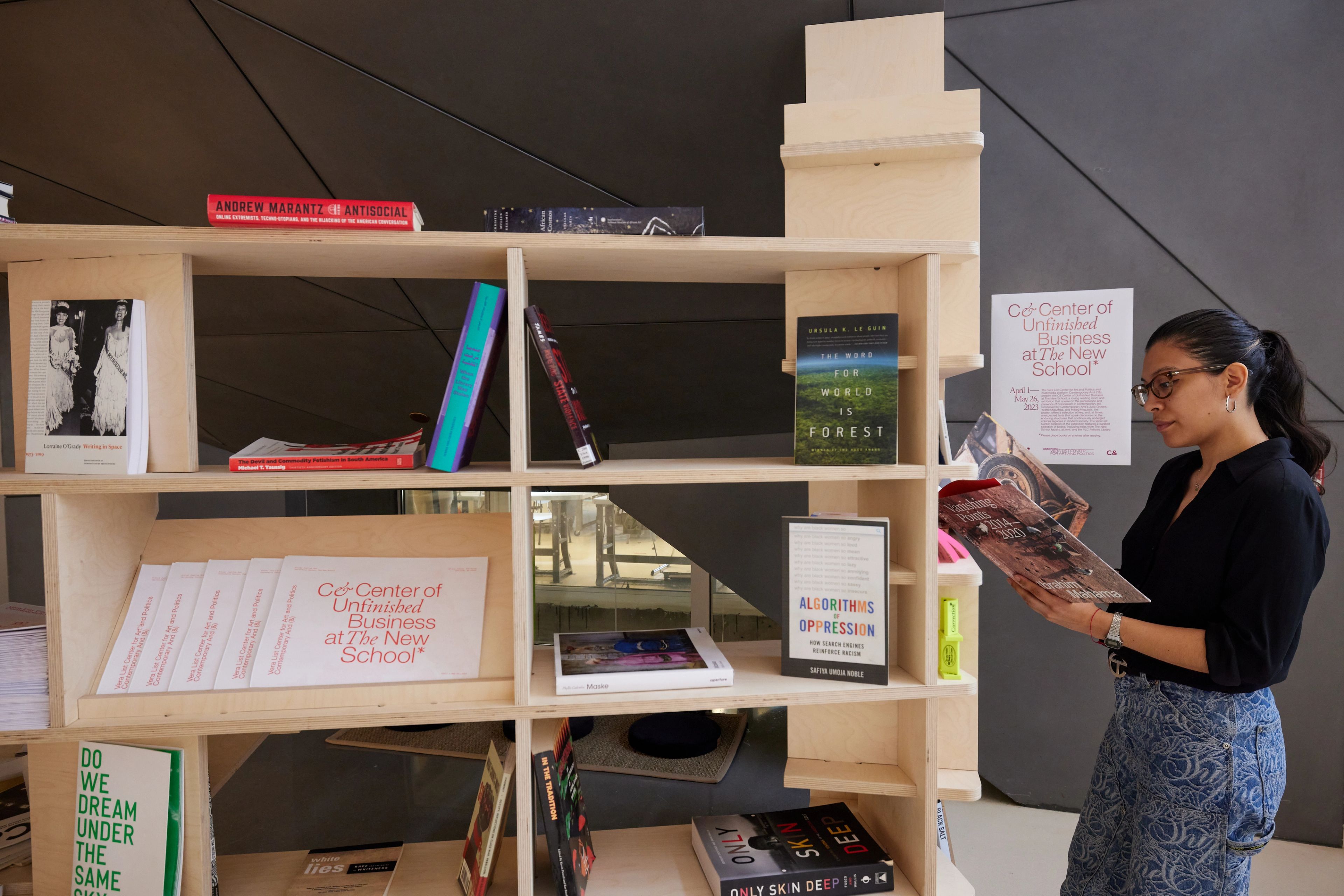 A woman reads a book next to a wooden bookshelf featuring books and materials for "Cd-Center of Unfinished Business at The New School."