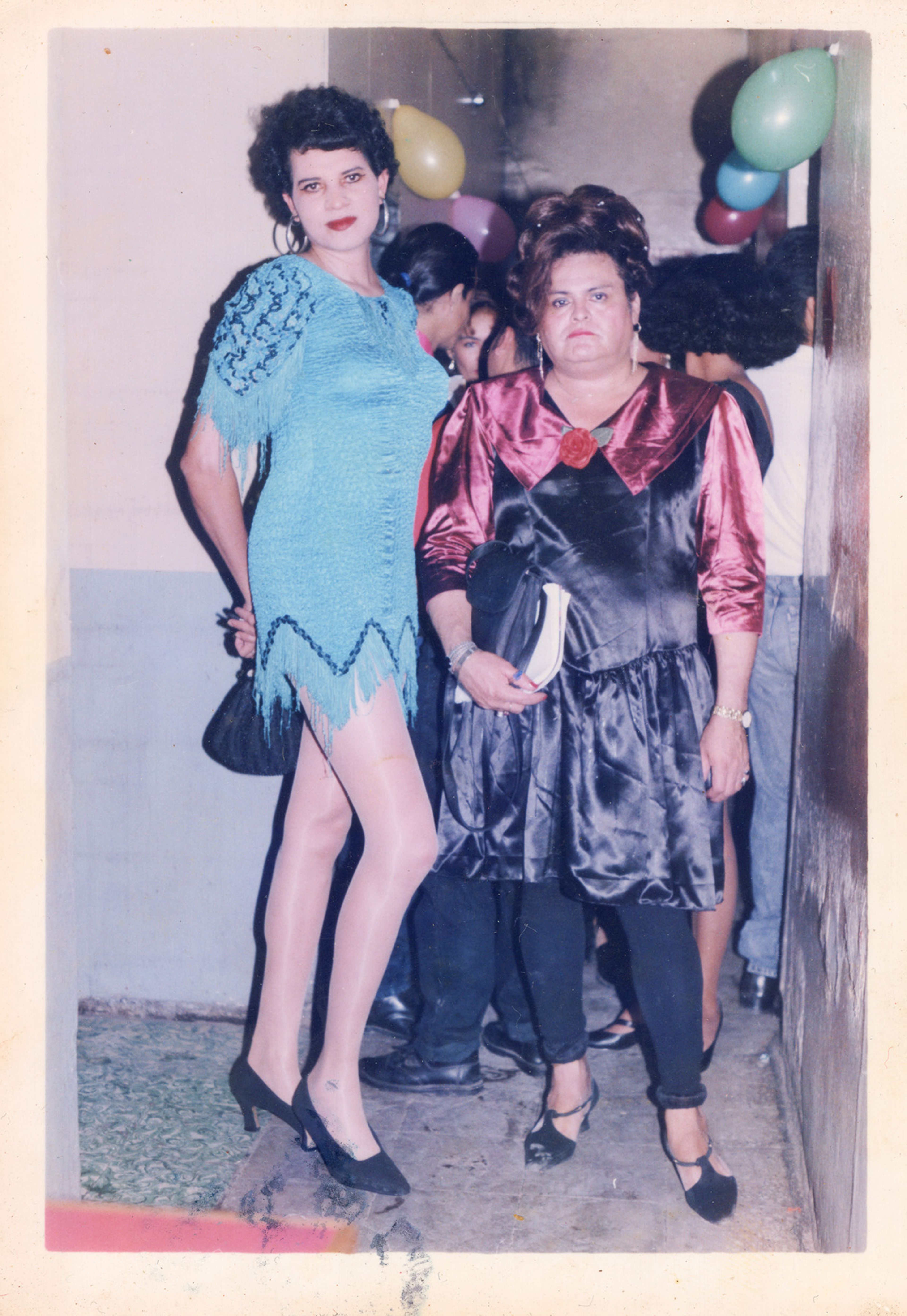 Two women in party attire stand in front of party balloons. The taller woman on the left wears a blue fringed dress, sheer hose, and heels. The shorter woman on the right wears a black dress with shiny maroon puffed sleeves and collar, black leggings, and T-strap heels.