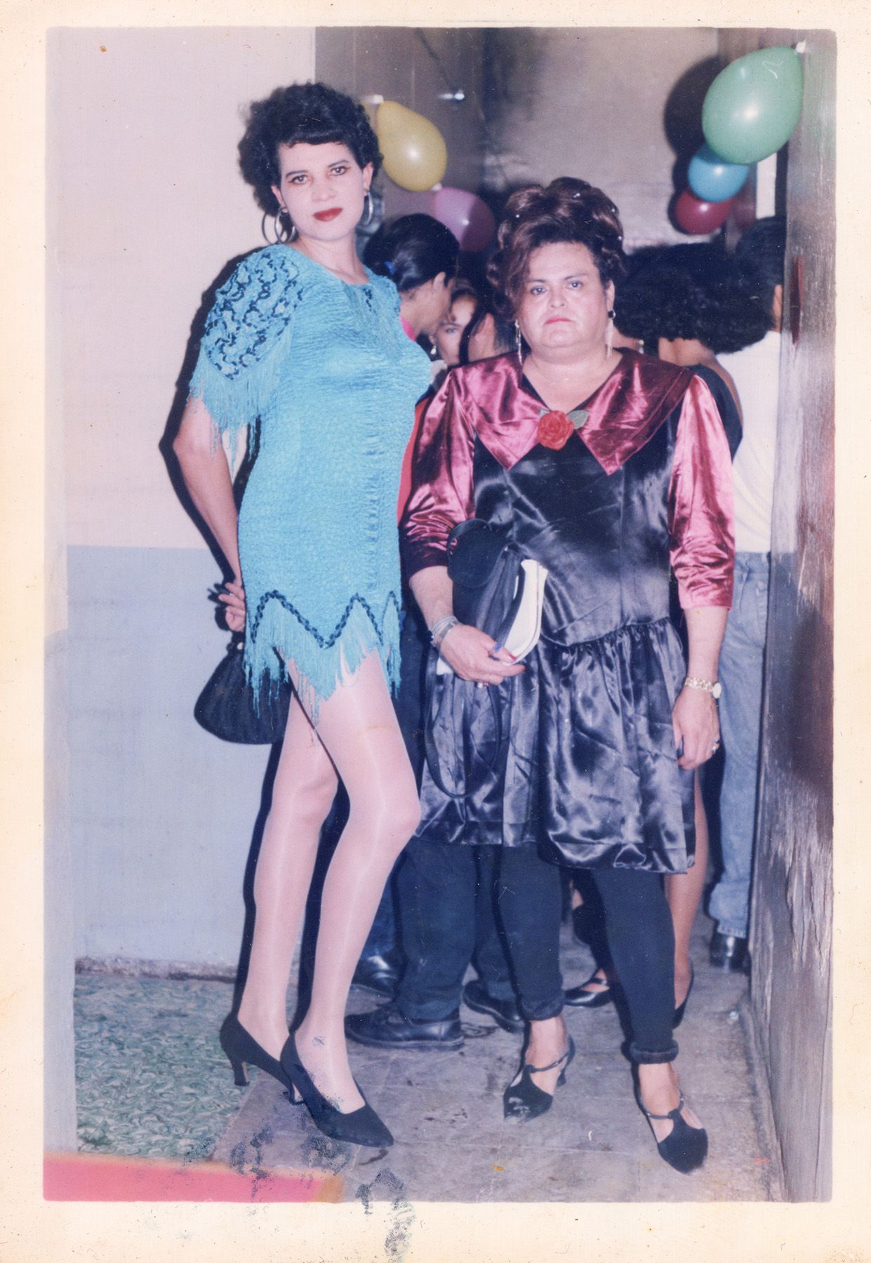 Two women in party attire stand in front of party balloons. The taller woman on the left wears a blue fringed dress, sheer hose, and heels. The shorter woman on the right wears a black dress with shiny maroon puffed sleeves and collar, black leggings, and T-strap heels.