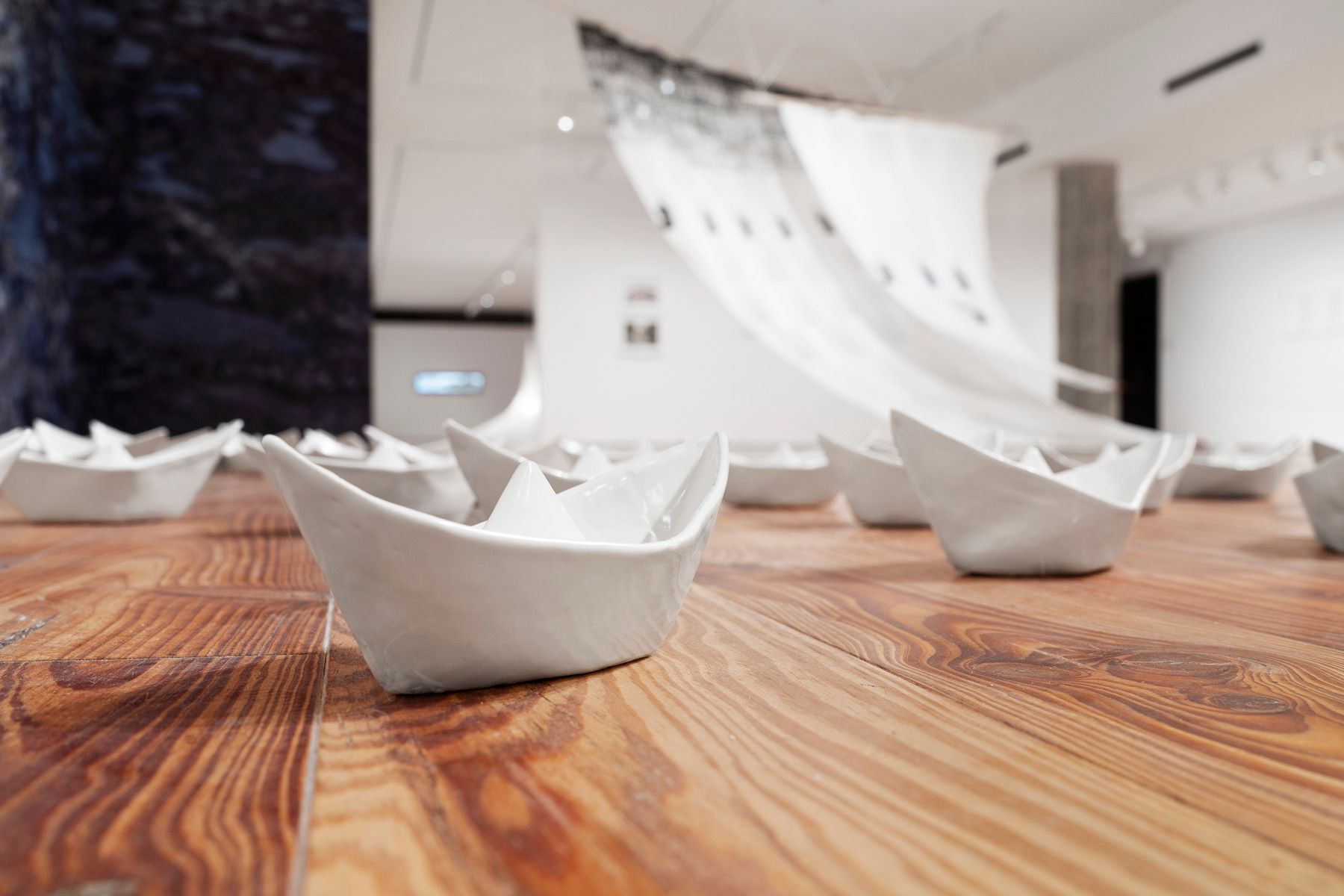 An art installation of white ceramic paper boats on a wooden floor, with a flowing fabric sculpture in the background.