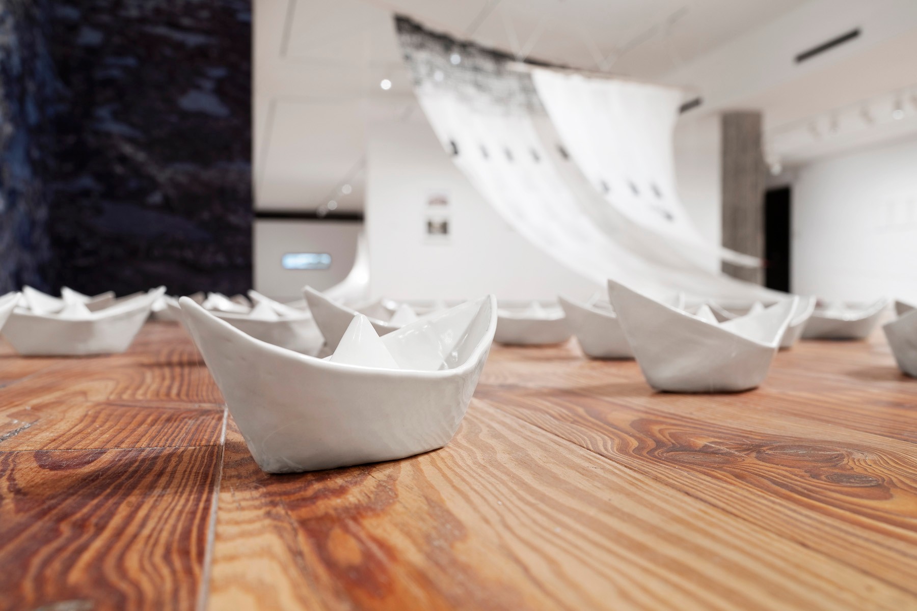An art installation of white ceramic paper boats on a wooden floor, with a flowing fabric sculpture in the background.