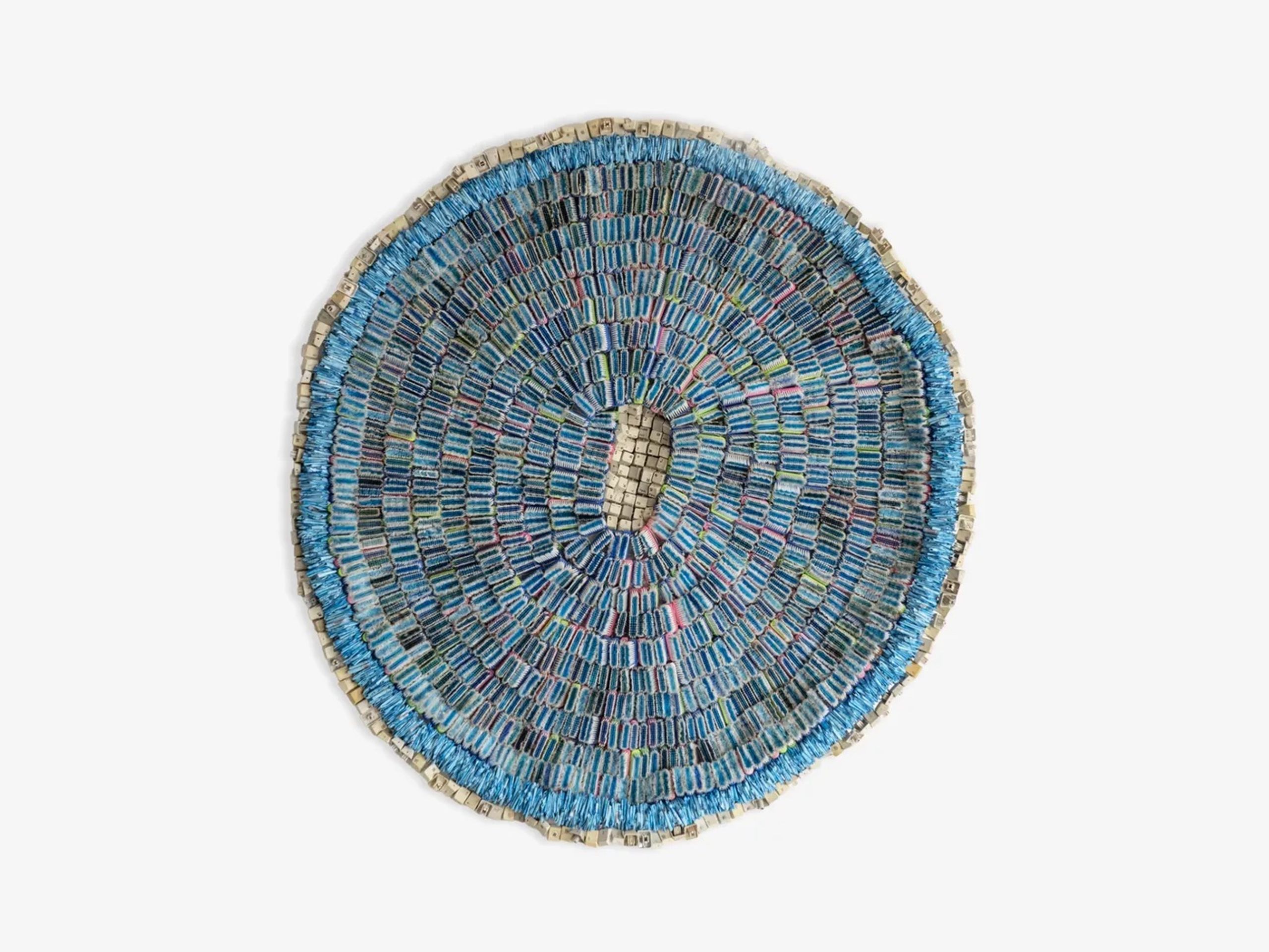Circular artwork made of small blue and grey rectangular pieces arranged in concentric rings, with a central oval opening.