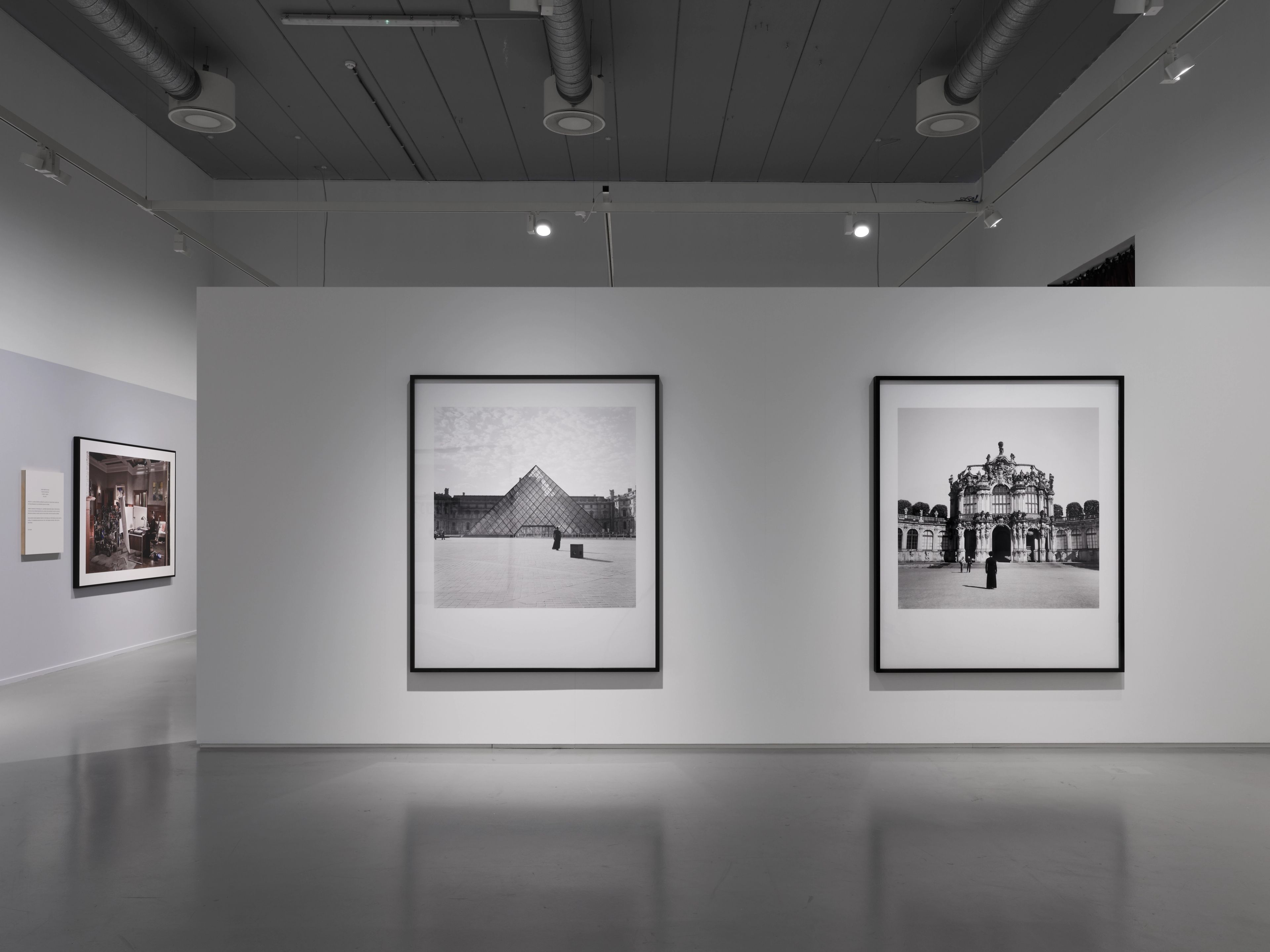 Art gallery with two large, framed black and white photos: one of the Louvre Pyramid, another of an ornate building.