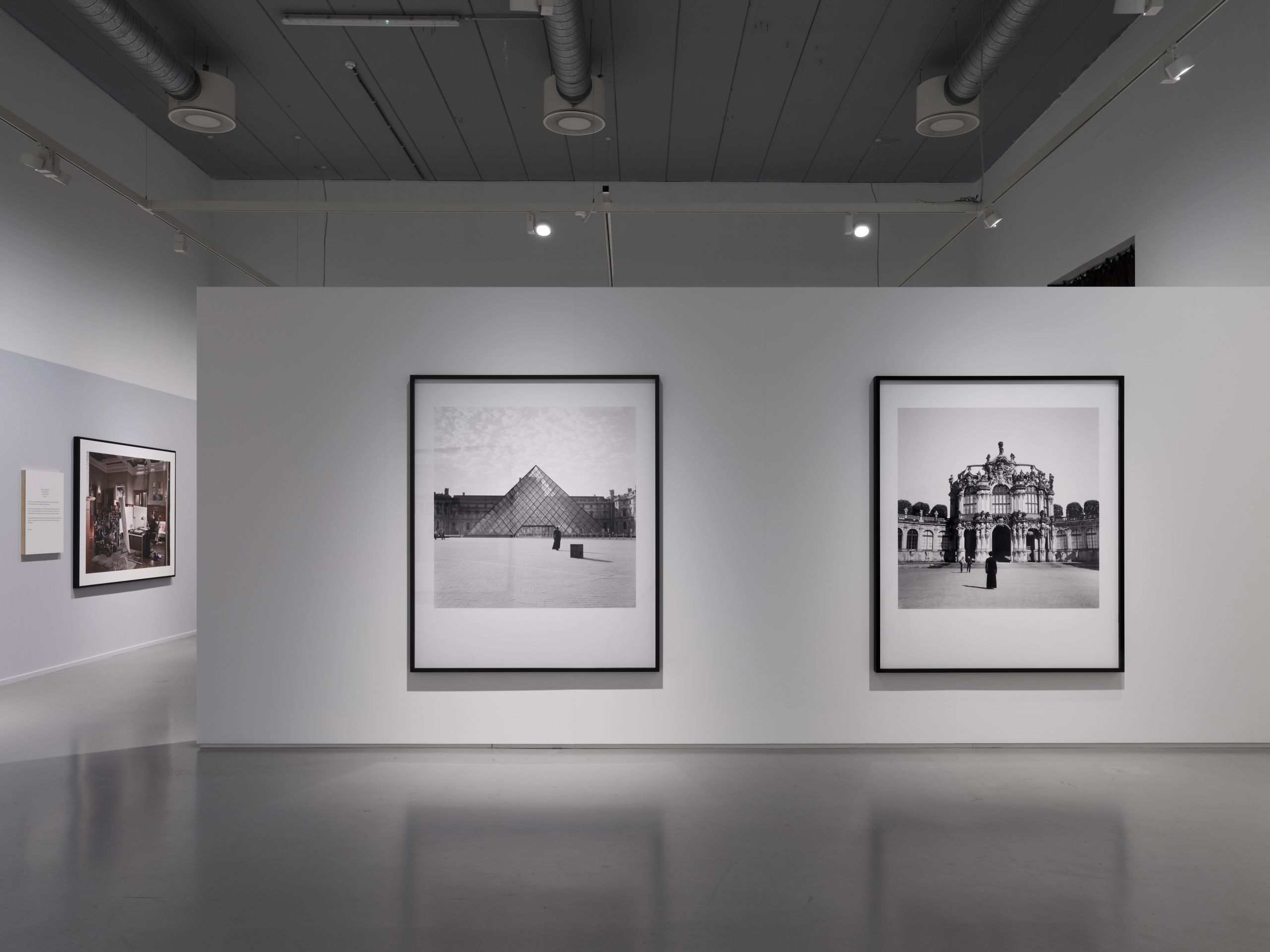 Art gallery with two large, framed black and white photos: one of the Louvre Pyramid, another of an ornate building.