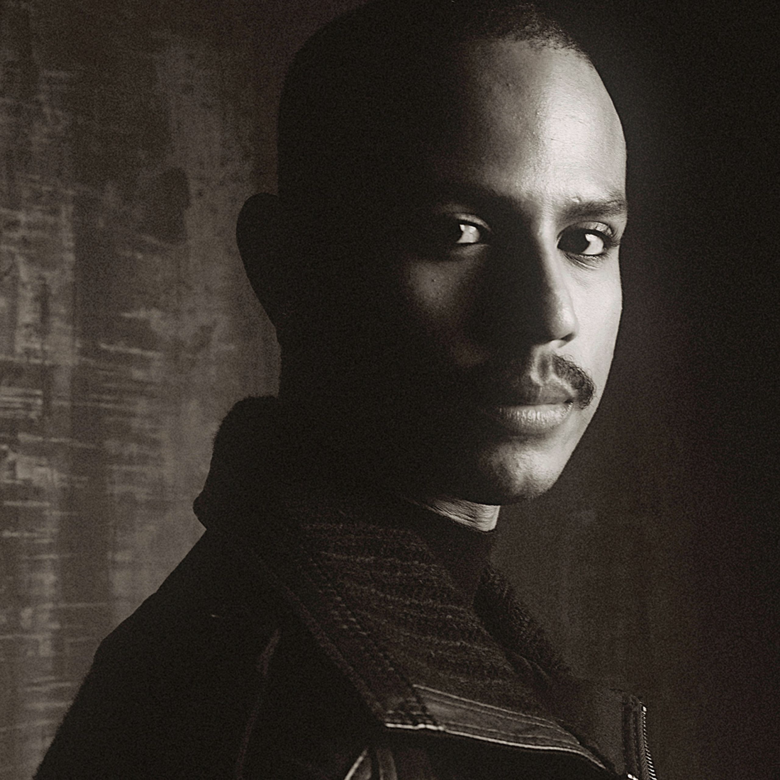 Sepia-toned, high-contrast portrait of a man with a mustache looking directly at the camera, his face partially in shadow.