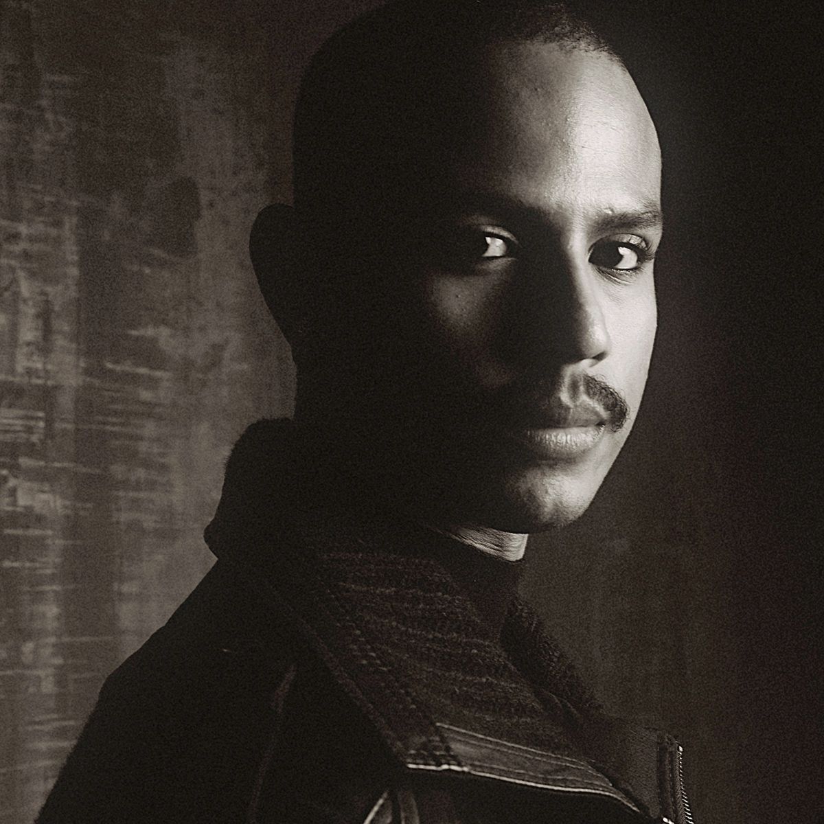 Sepia-toned, high-contrast portrait of a man with a mustache looking directly at the camera, his face partially in shadow.
