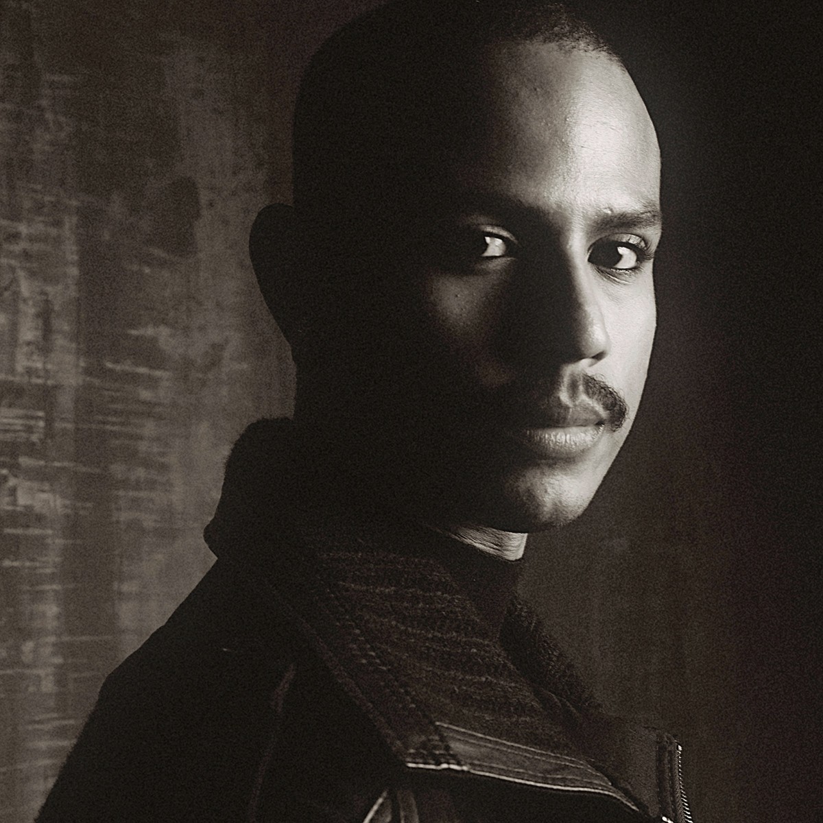 Sepia-toned, high-contrast portrait of a man with a mustache looking directly at the camera, his face partially in shadow.