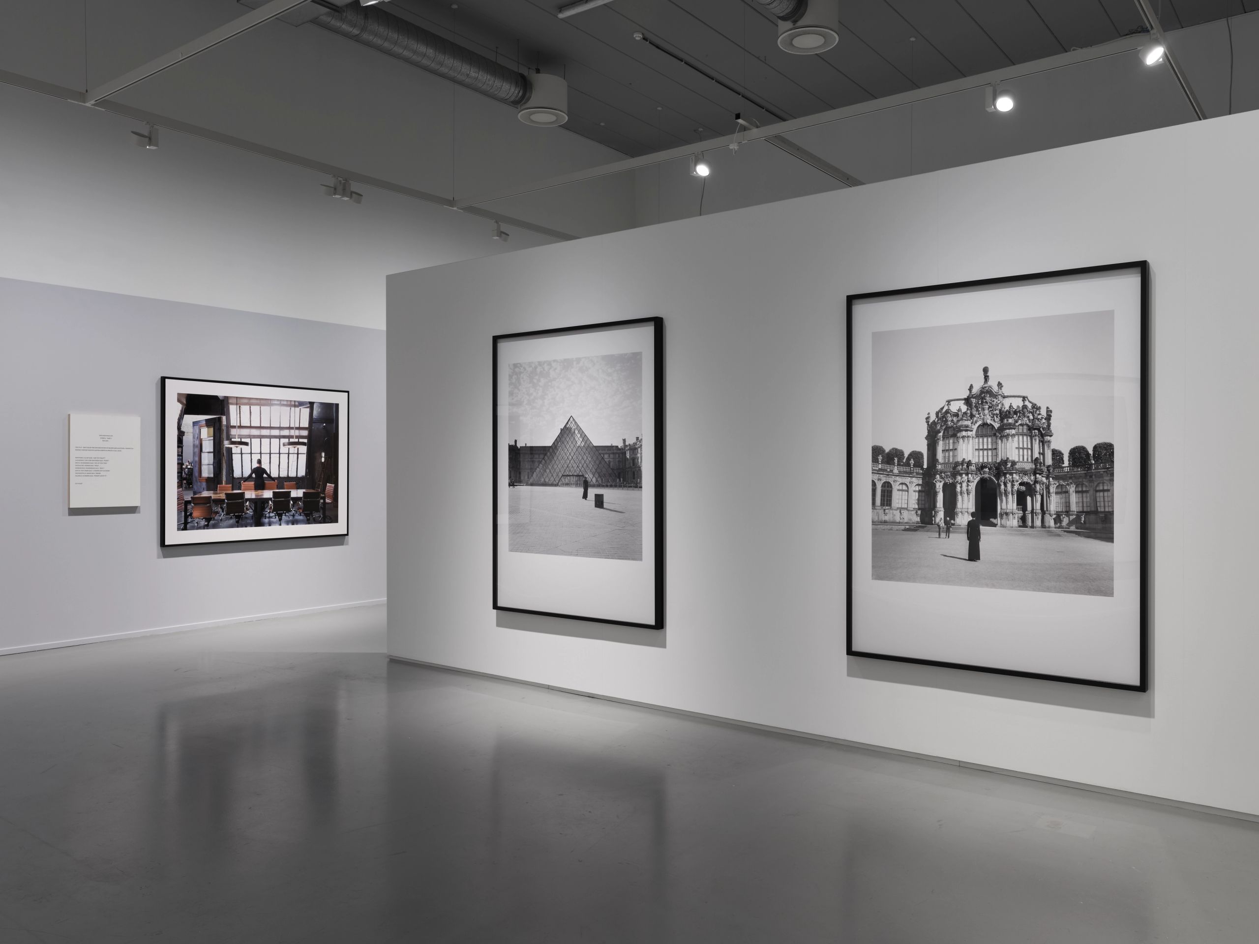 A modern art gallery with white walls and a gray floor, featuring two large black-and-white architectural photographs on the right and a color interior photograph on the left.