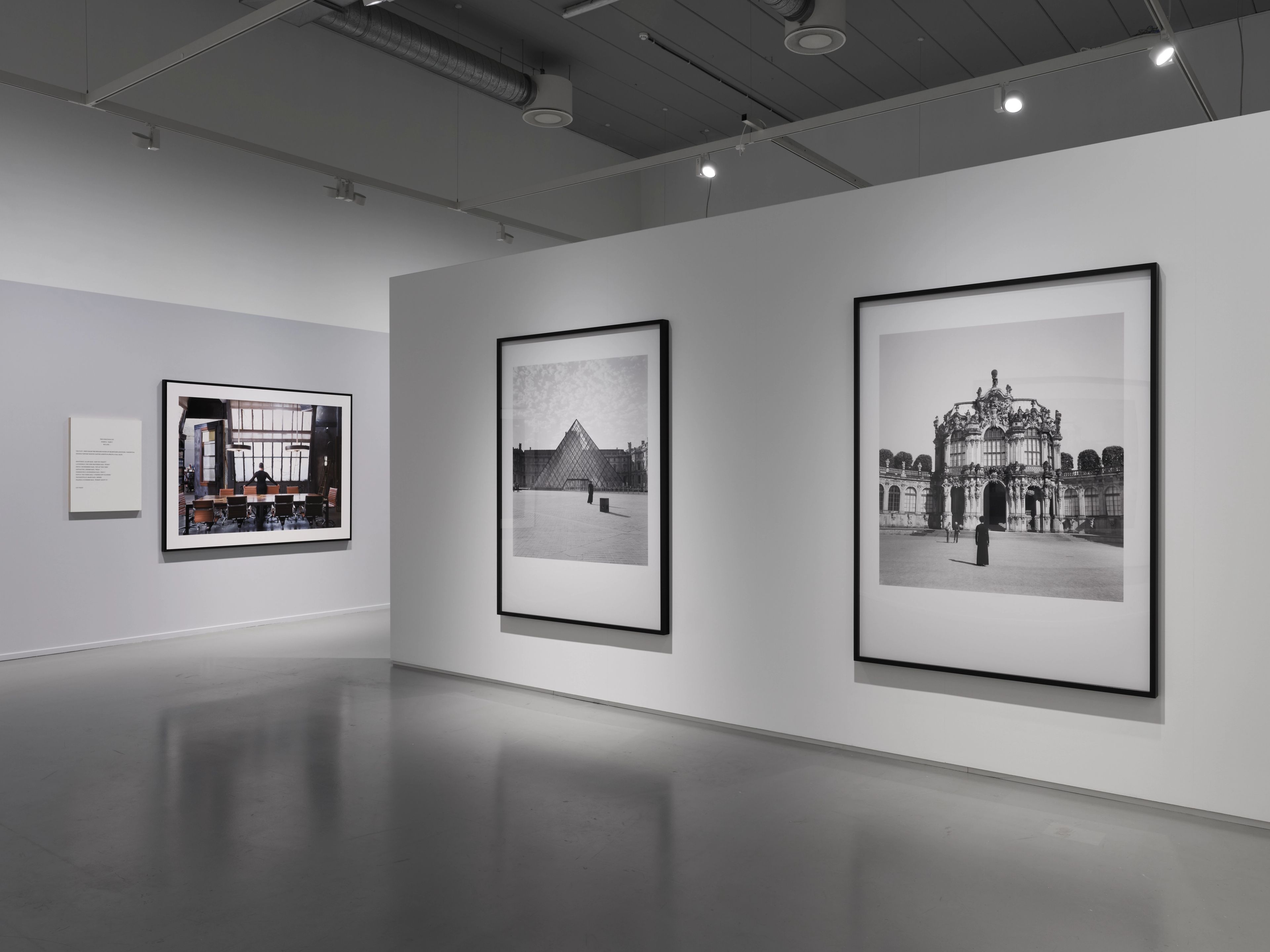 A modern art gallery with white walls and a gray floor, featuring two large black-and-white architectural photographs on the right and a color interior photograph on the left.