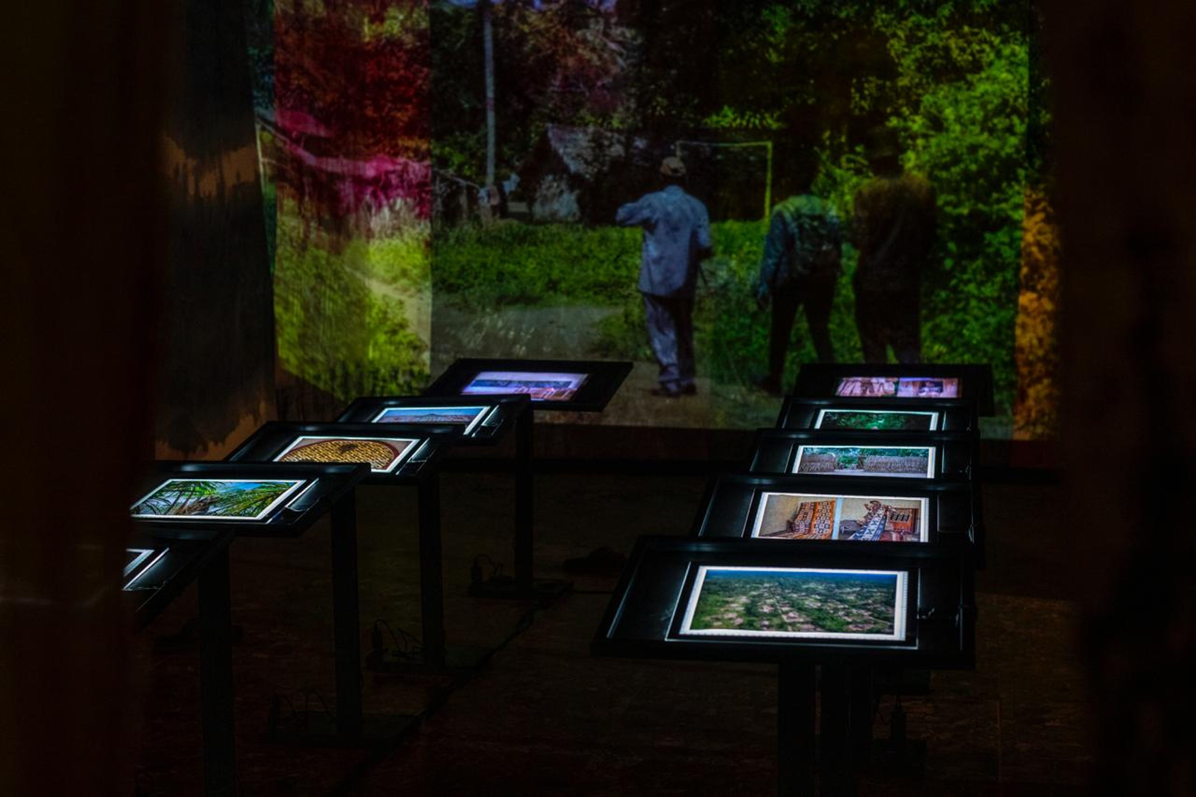 Multimedia exhibit with a large projected image of people outdoors and several illuminated screens on stands in a dark room.