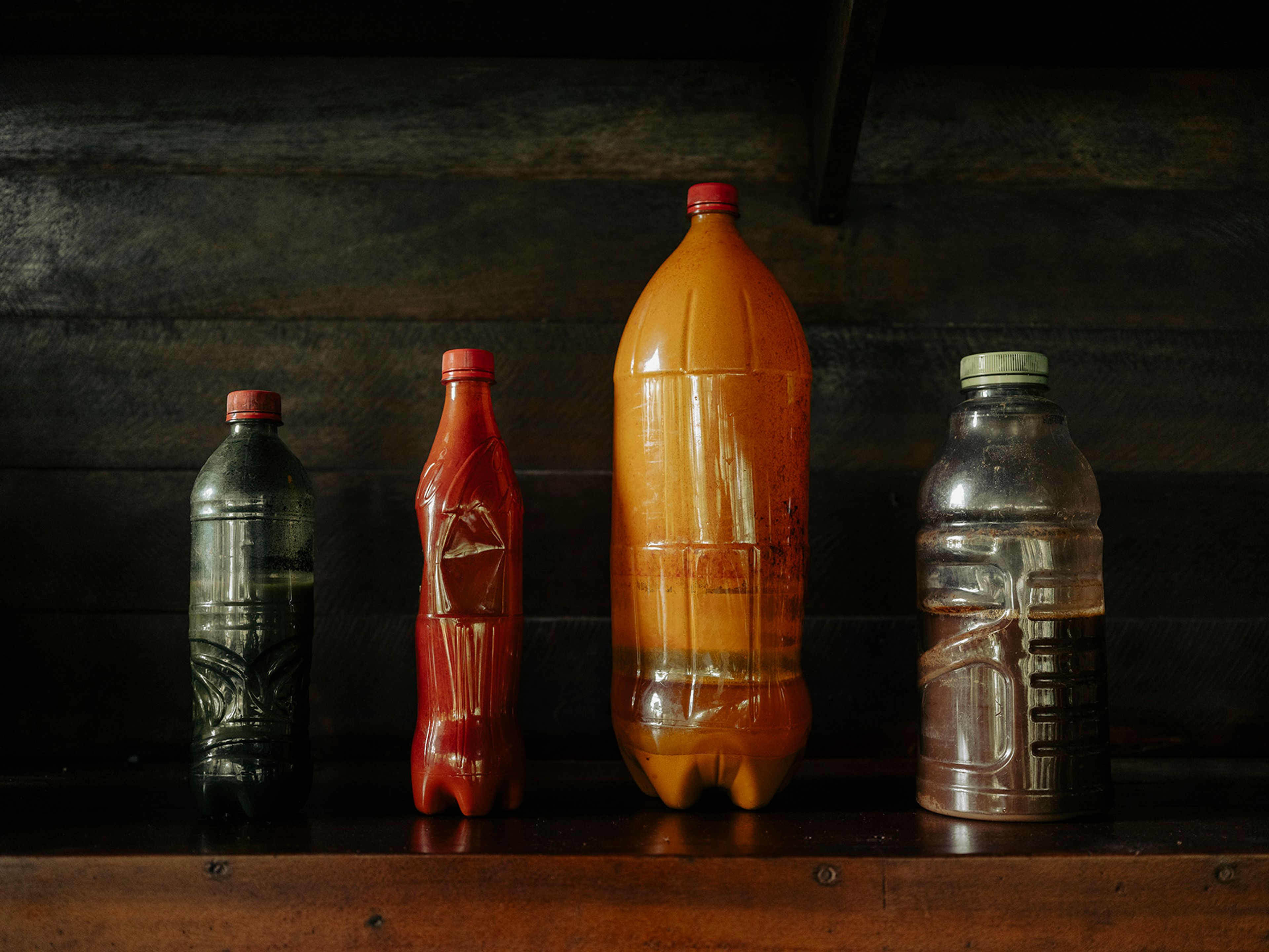 Four plastic bottles of varying sizes and colors, filled with different liquids, sit on a wooden shelf.