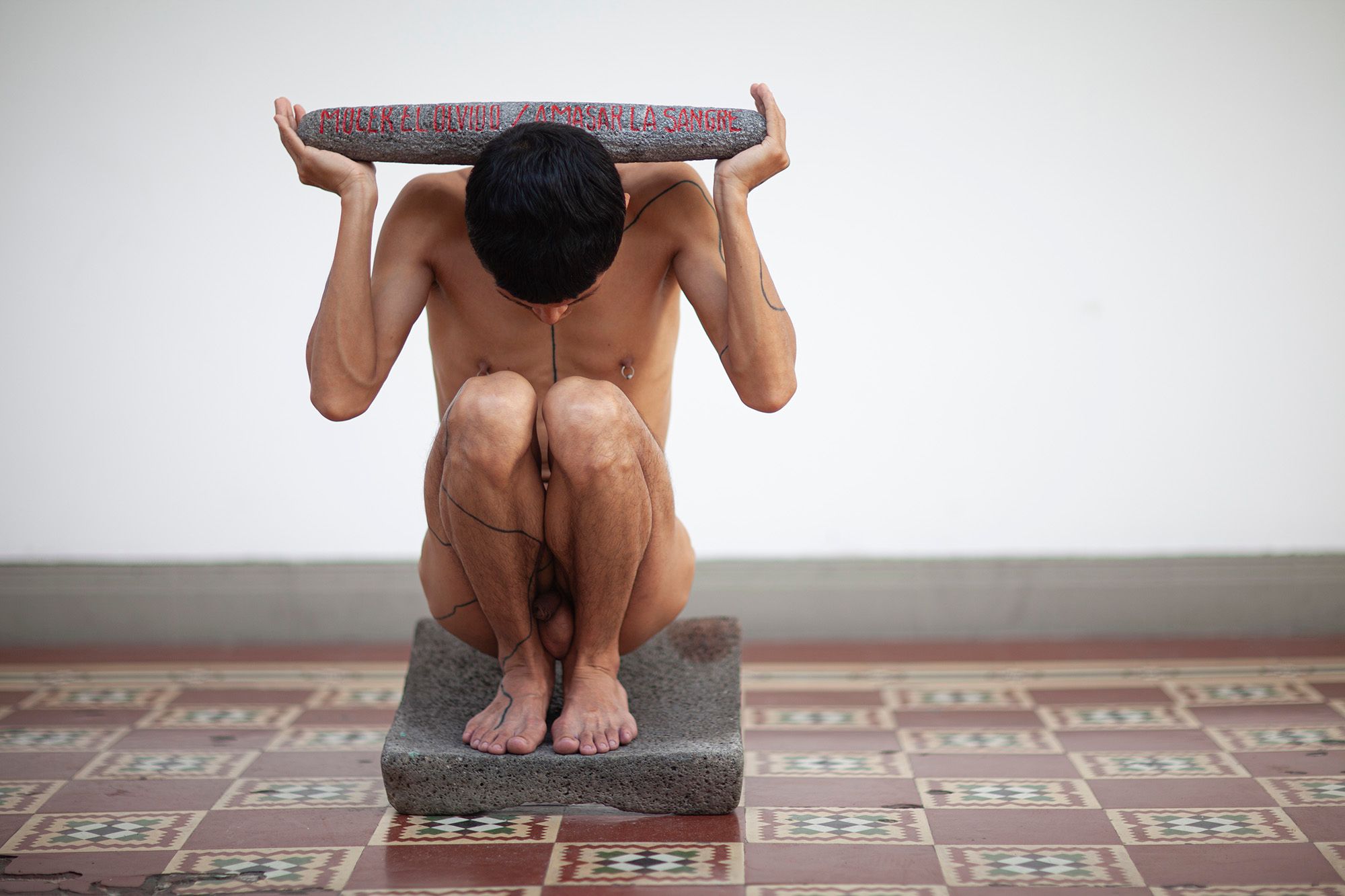 A person with dark lines on their body squats on a stone block, holding another stone block with red carved text above their head.