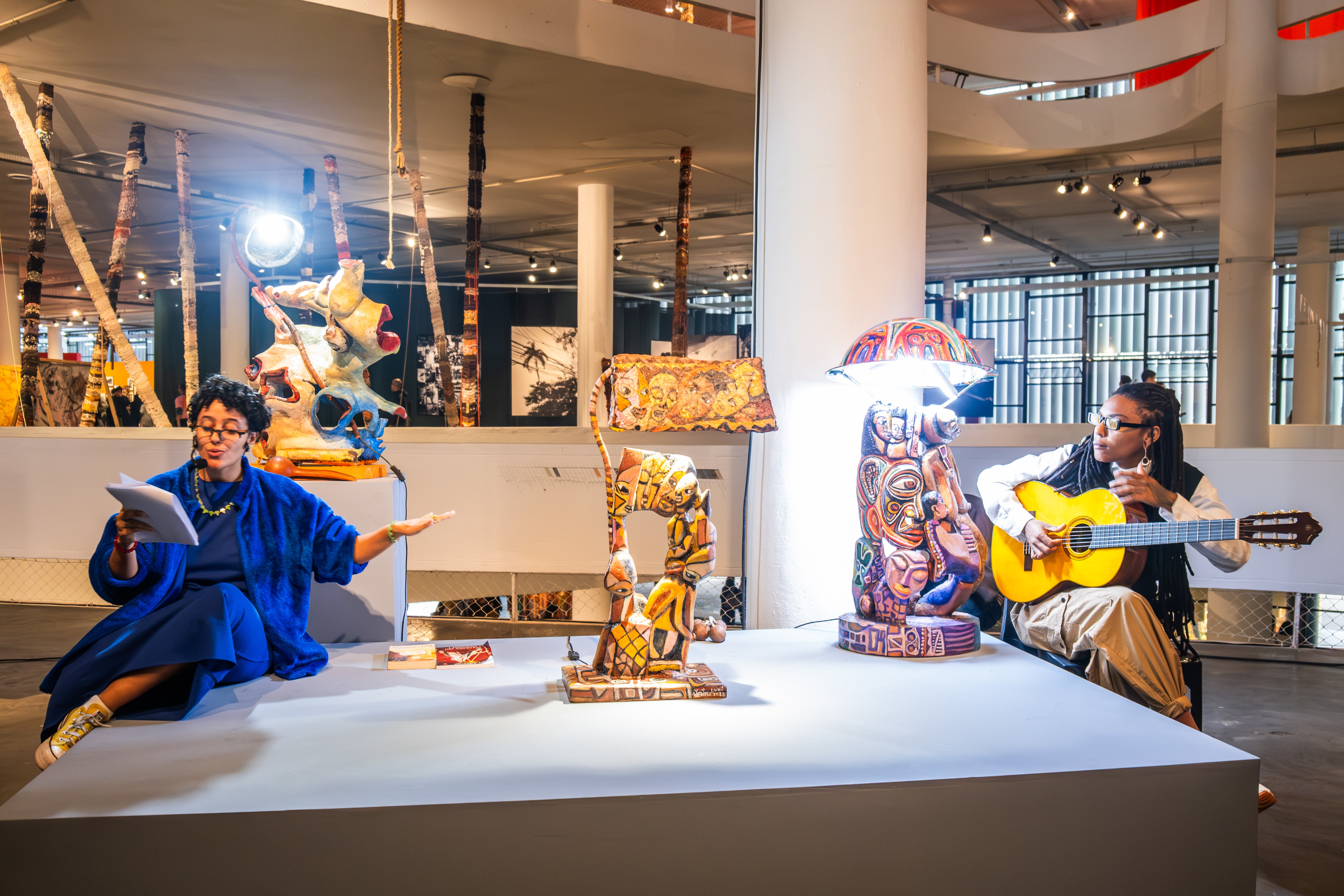 A woman in blue recites while another plays guitar, surrounded by colorful sculptures in a gallery.