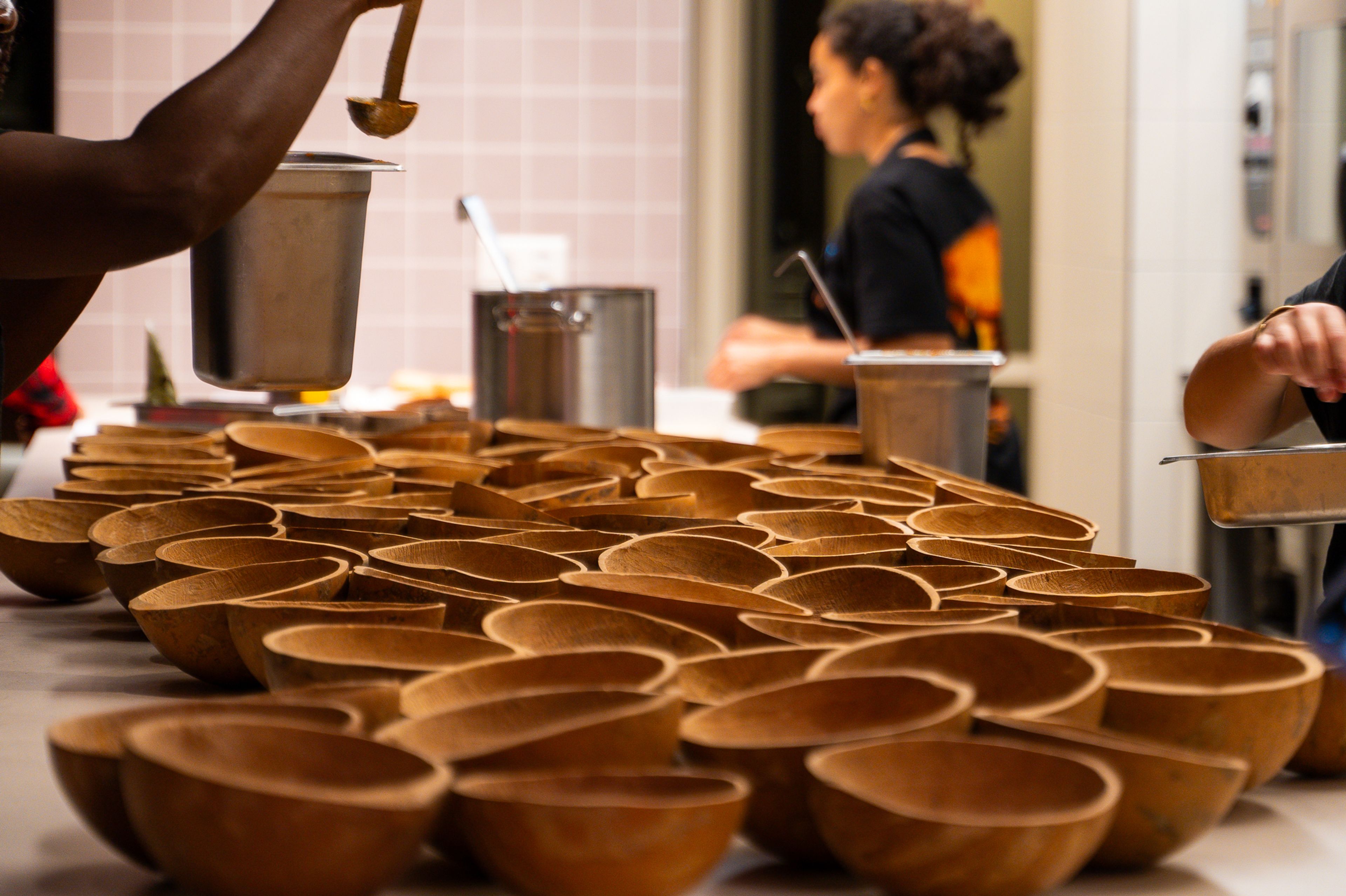 Rows of empty wooden bowls are set on a table, with people preparing food in the blurred background.