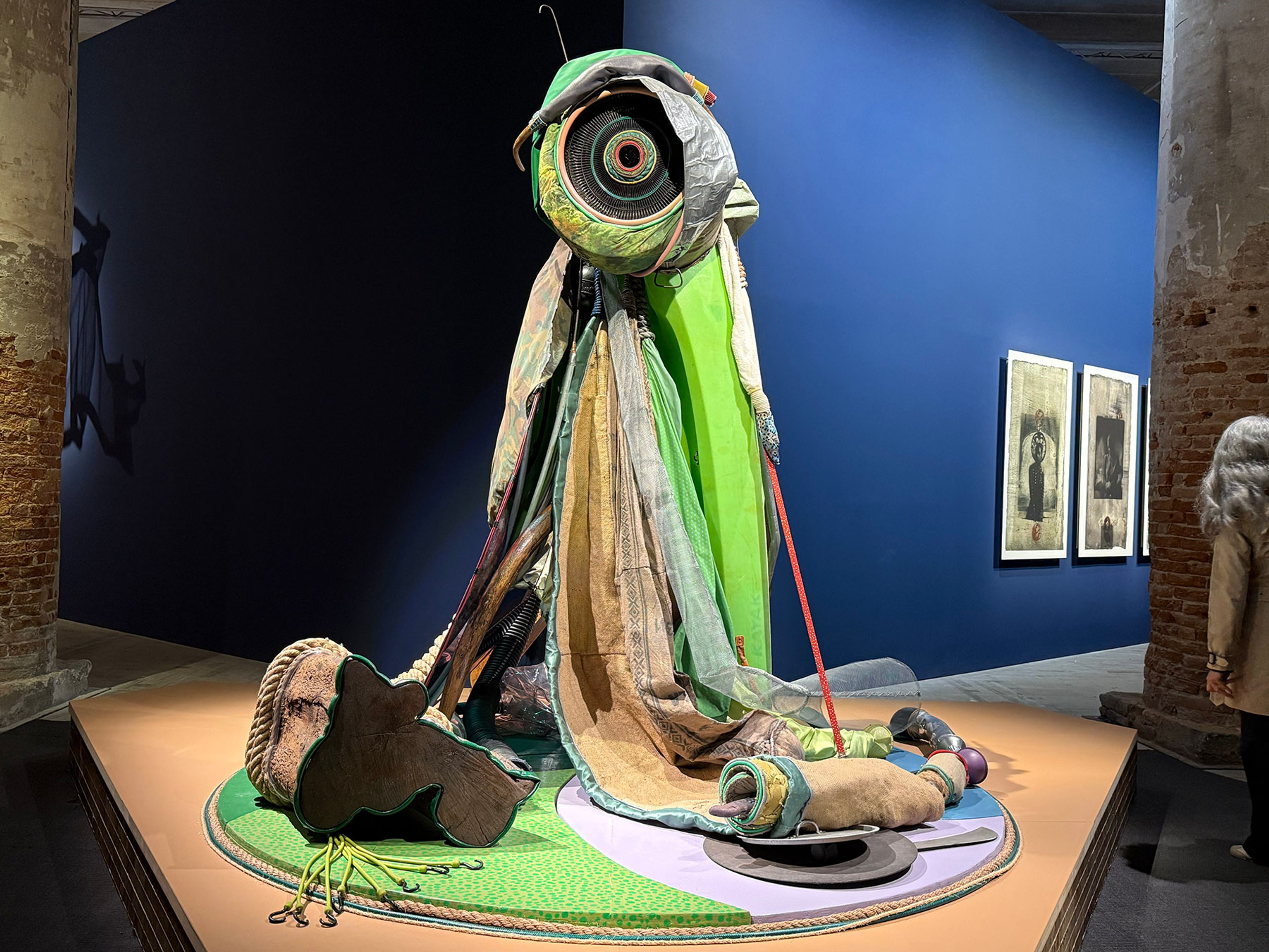 A large, colorful abstract sculpture made of draped textiles with a circular patterned top, displayed on a round green base in an art gallery.