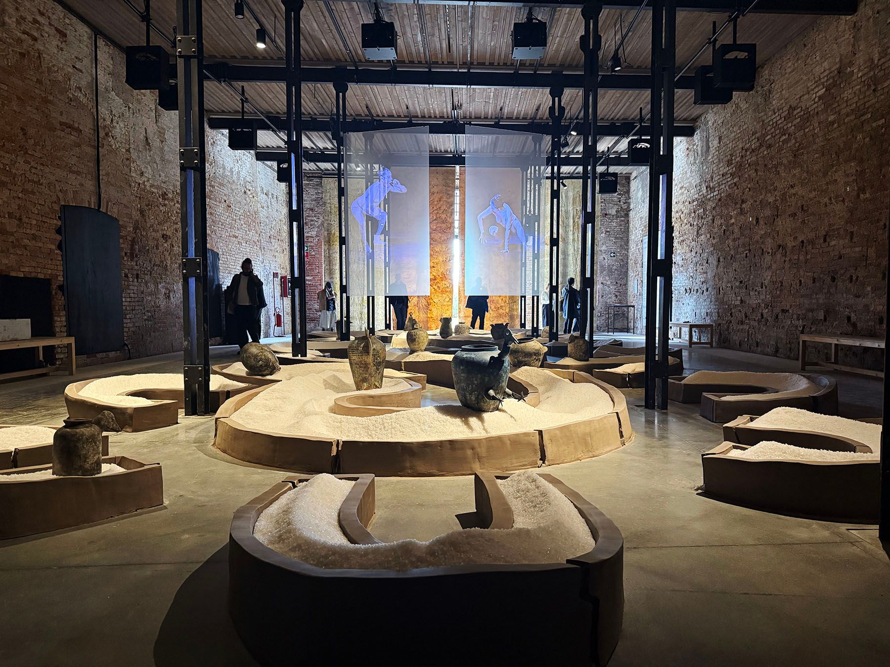 Art exhibition in a brick-walled room with curved displays of white material showcasing ancient vessels and sculptures. Abstract blue figures are projected on background screens.