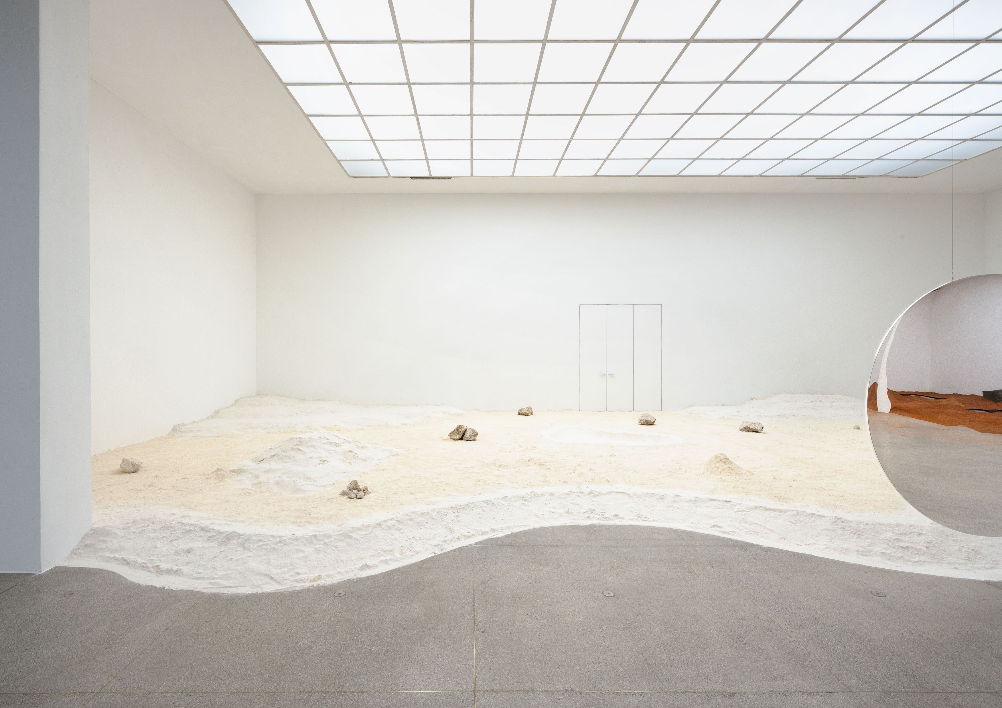 A gallery with a grid skylight shows a wavy sand and rock landscape, bordered by concrete, with a large reflective sphere mirroring a warm interior on the right.