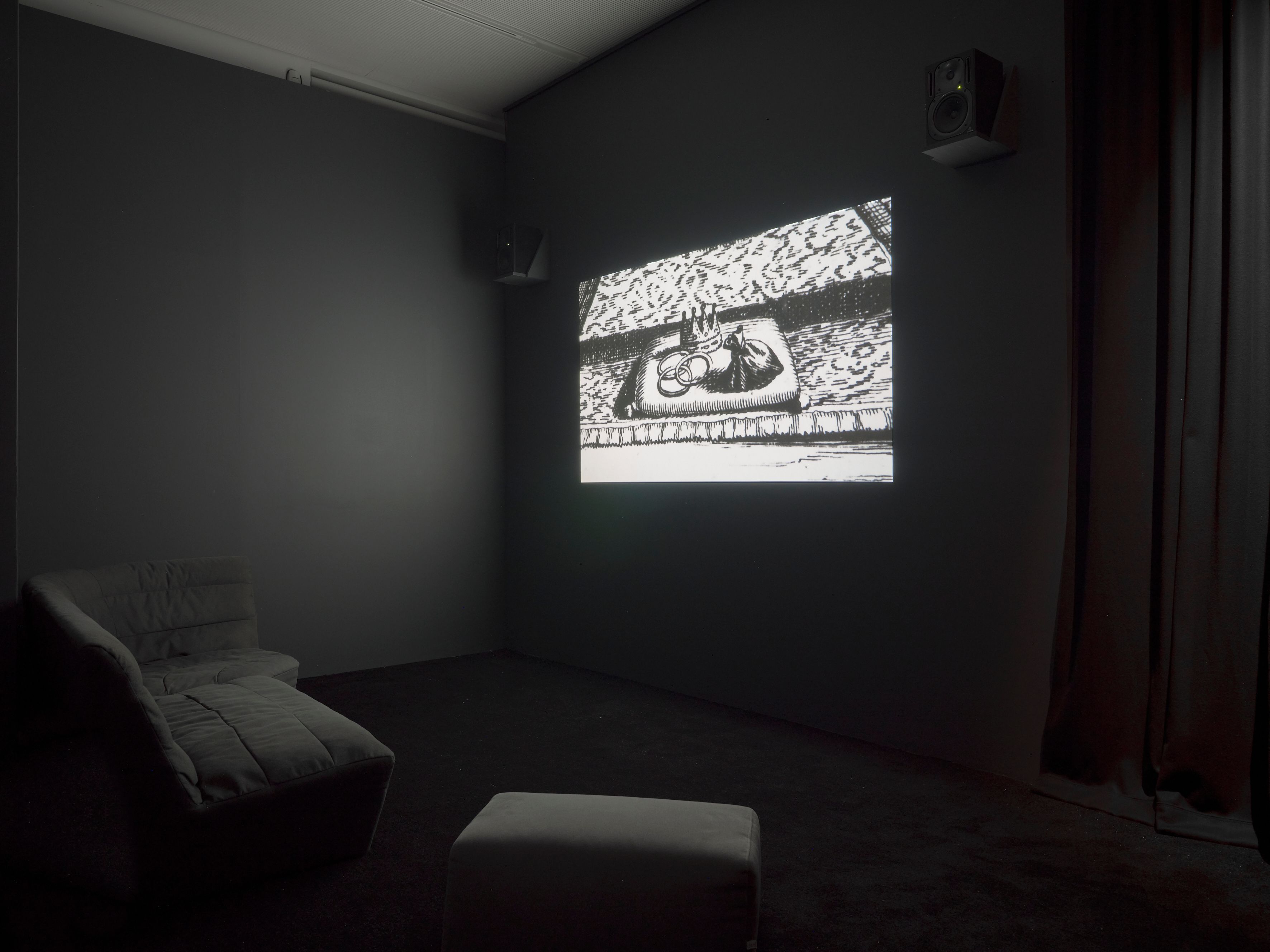 A dark room features an ornate black-and-white image projected on a wall, flanked by speakers, with lounge seating in the foreground.